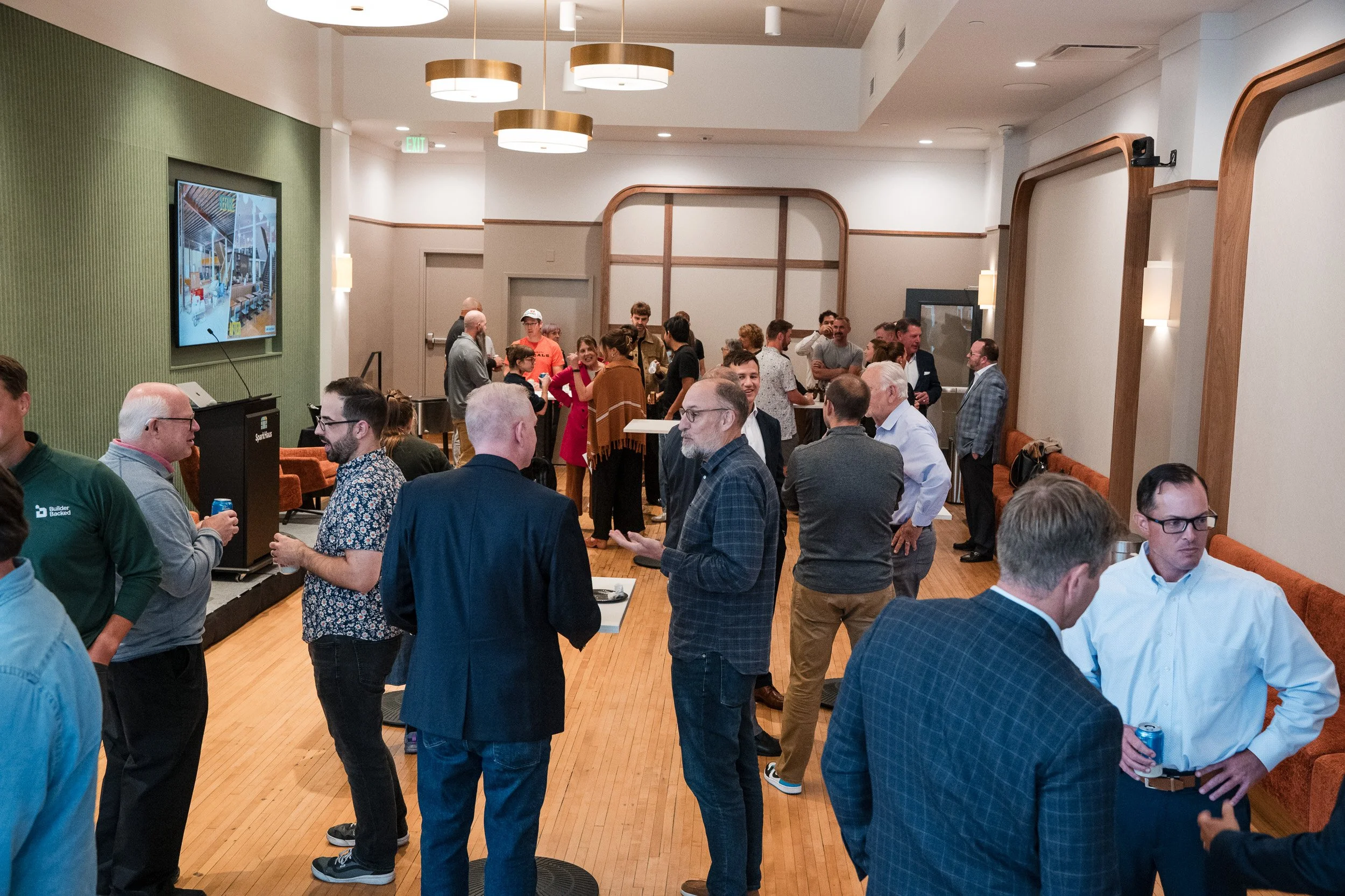 A group of people gathered in a modern, well-lit room with wooden flooring and green and beige walls, engaging in conversations during an event or networking session.