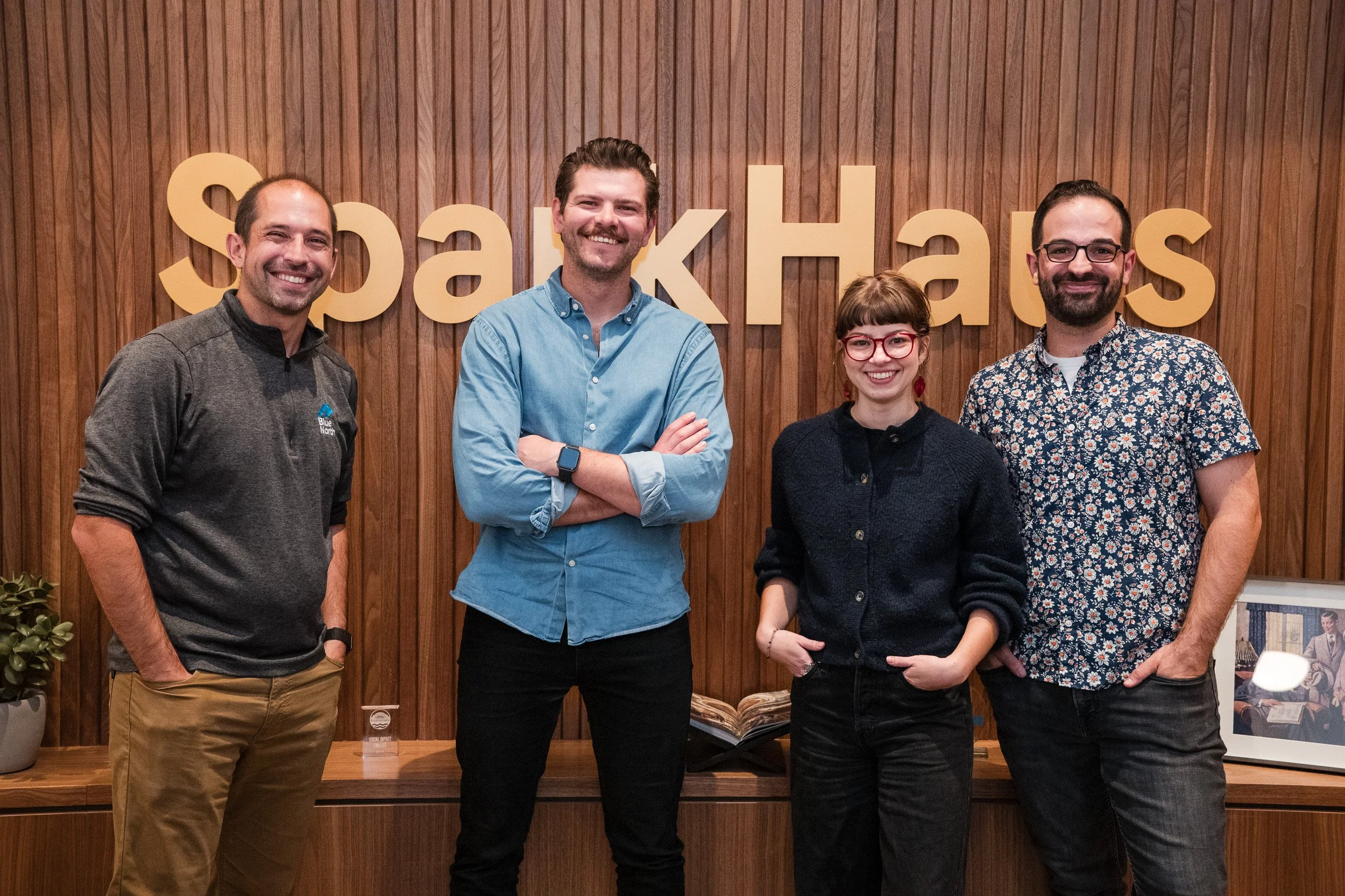 Four smiling people standing in front of a wooden wall with the word 'SparkHacks' in large letters.