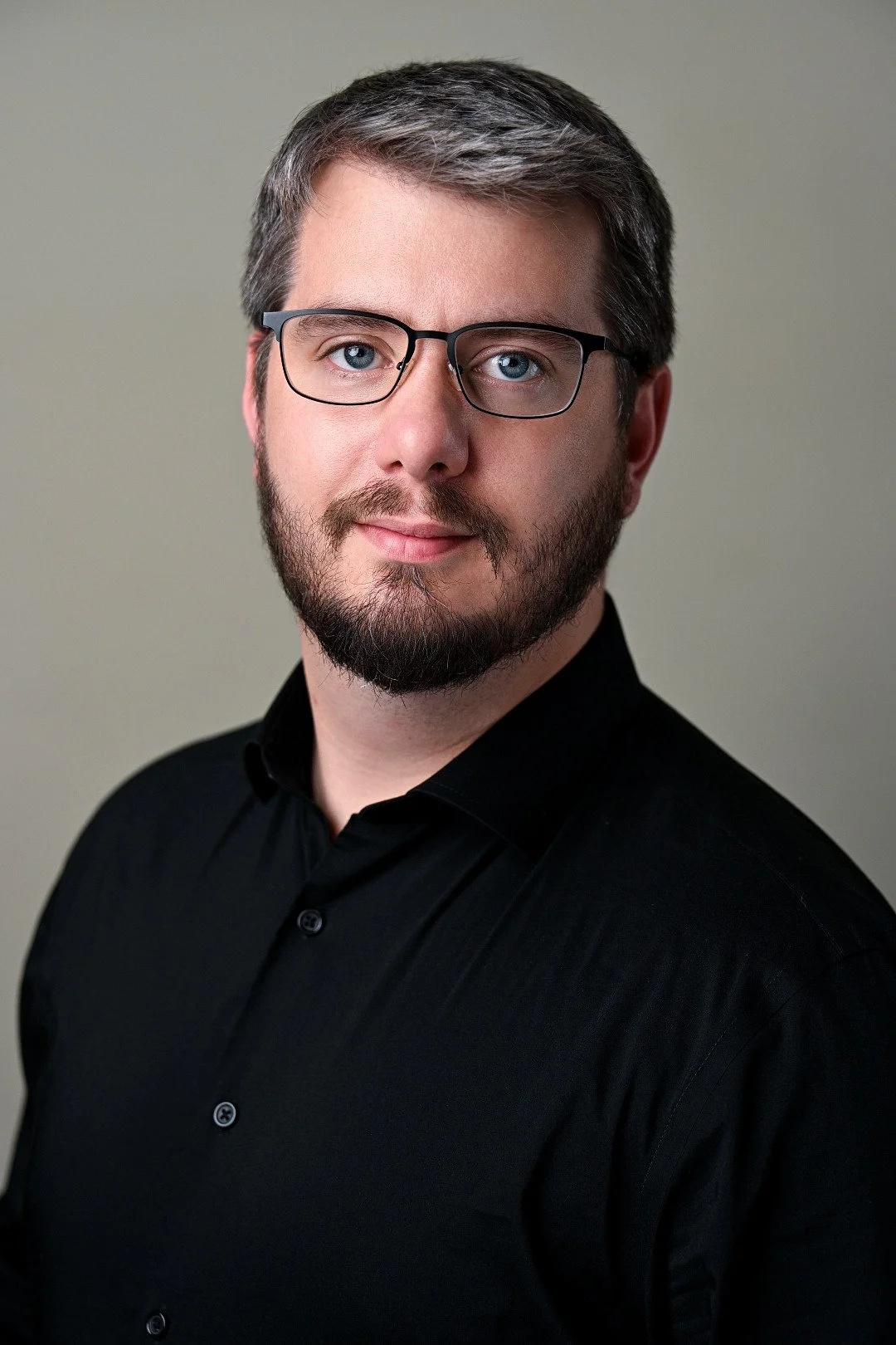 A man wearing glasses with a beard and mustache, in a black collared shirt, looking at the camera with a neutral expression against a plain neutral background.
