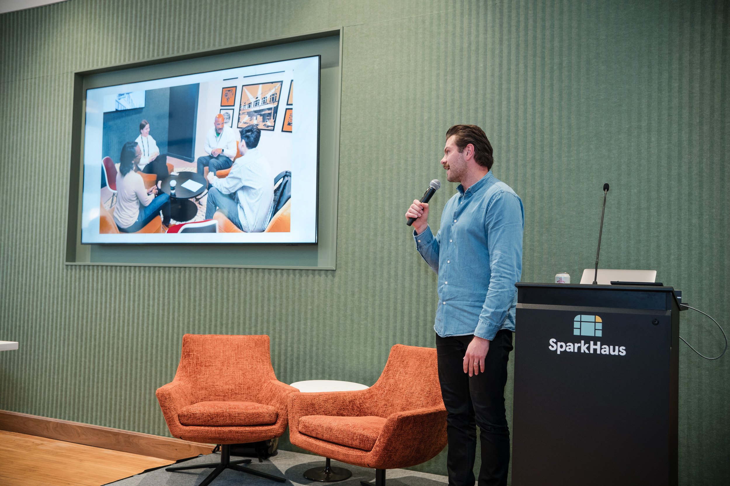 A man with dark hair and a beard, wearing a blue button-up shirt, stands holding a microphone in front of a large screen displaying a photo of four people sitting in a circle in a room with orange chairs. The room has green walls, a black podium with