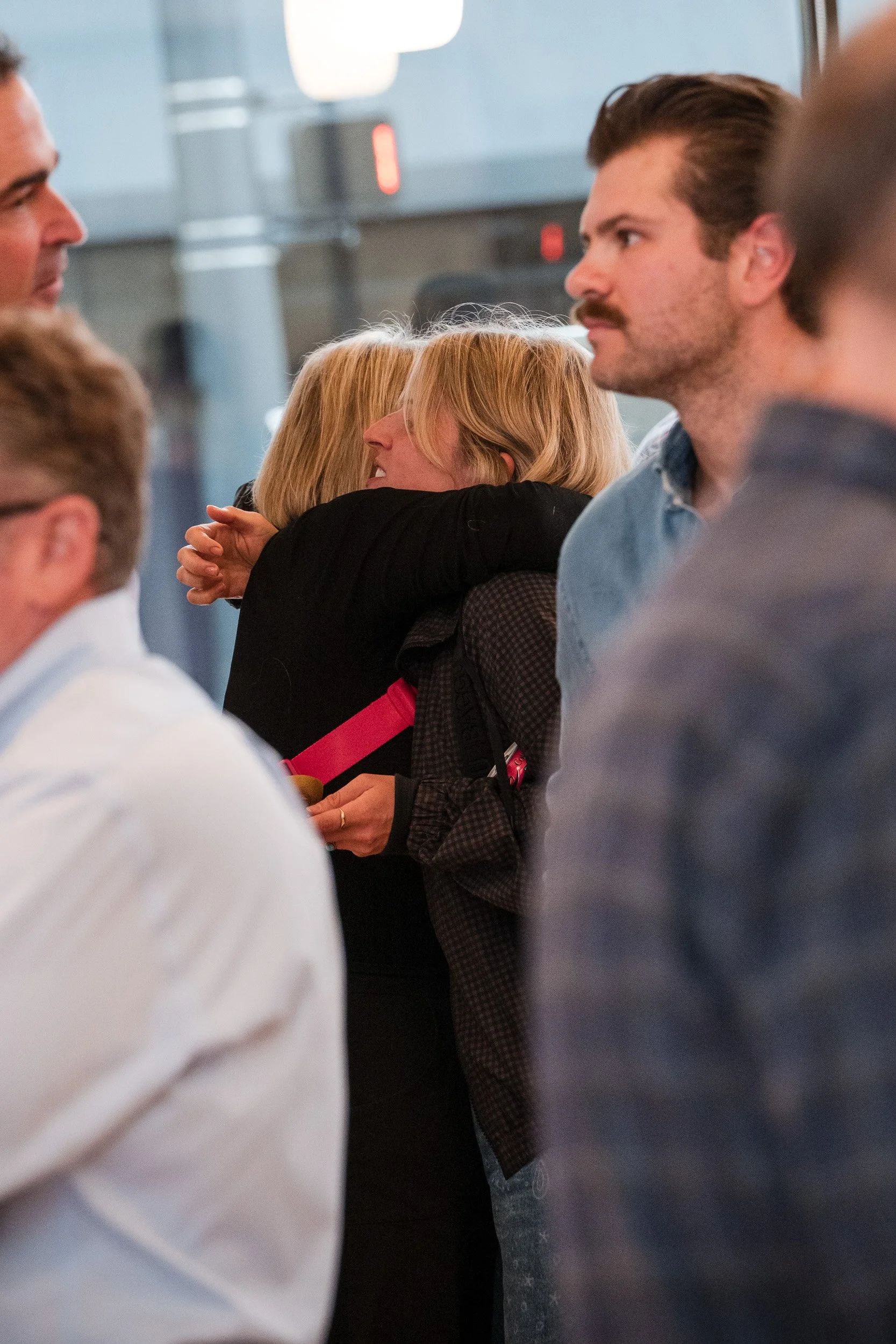 People in a crowded indoor setting, with two women hugging and several men standing around.