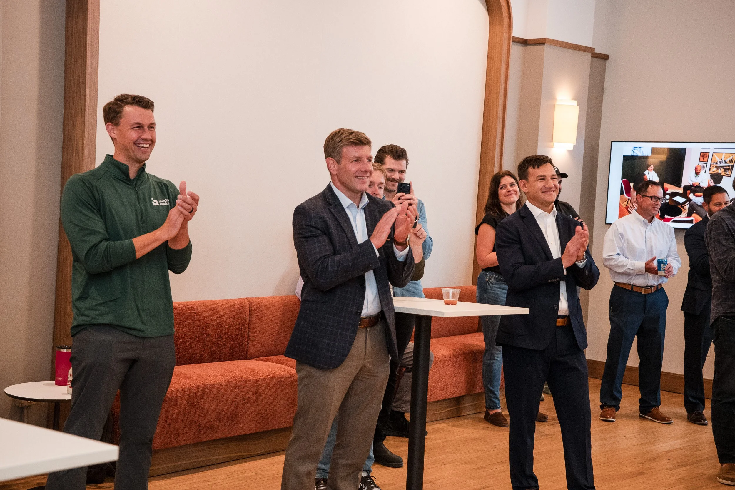 Group of people standing in a room, smiling and clapping, during a presentation or event. One person taking a photo, others holding drinks, and a large monitor displaying a video call or virtual meeting