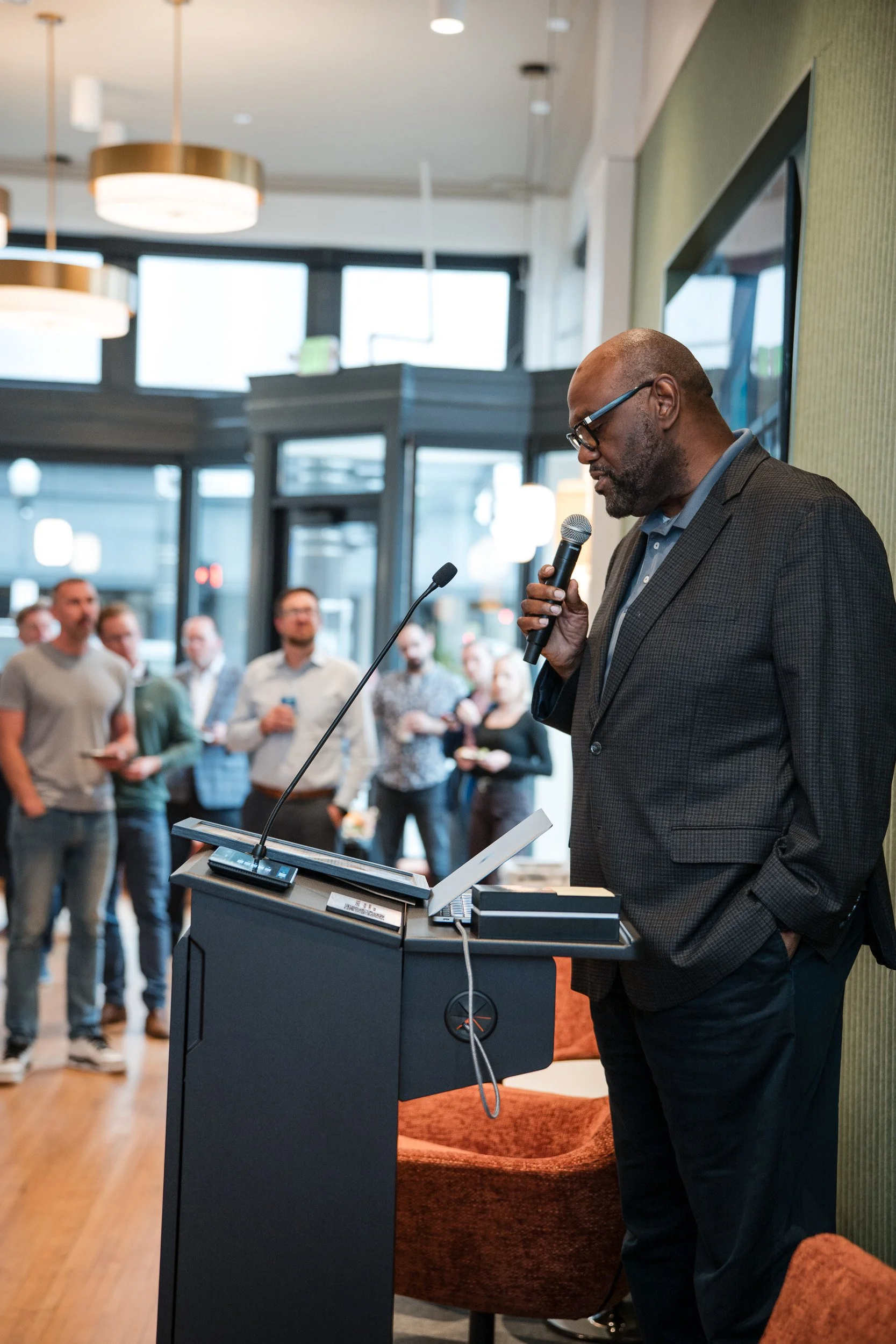 A man in a dark suit, wearing glasses, holding a microphone, and speaking at a podium inside a modern conference room or event space. Several people are standing in the background, listening.