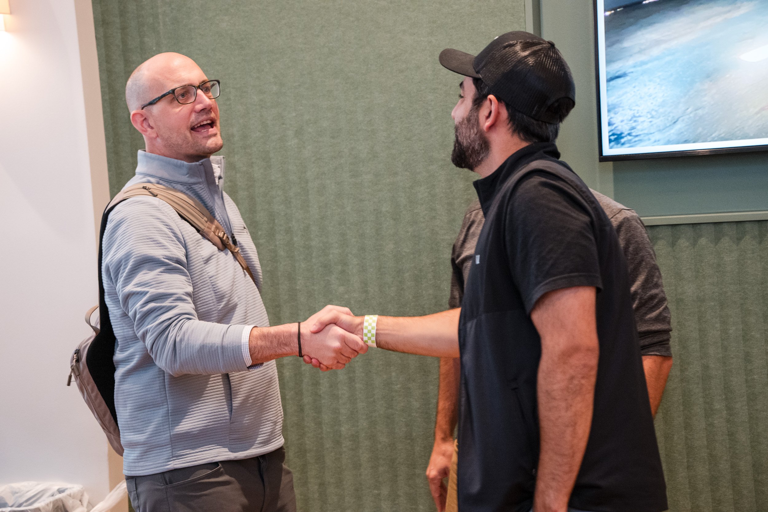 Two men shaking hands indoors, one with a backpack and glasses, the other wearing a black cap and black shirt.