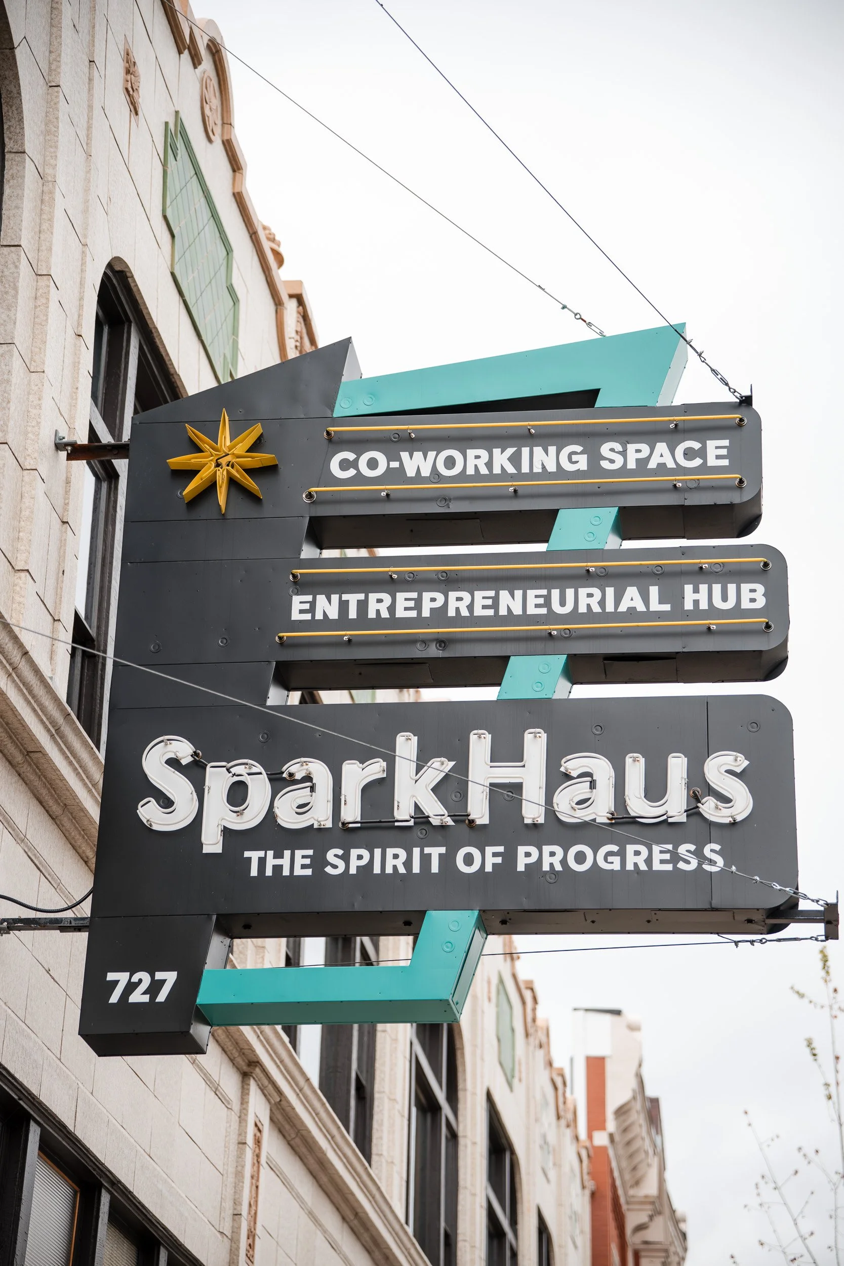 Street view of a signboard hanging on a building, reading 'CO-WORKING SPACE' and 'ENTREPRENEURIAL HUB' with a star logo, with other building facades visible in the background.