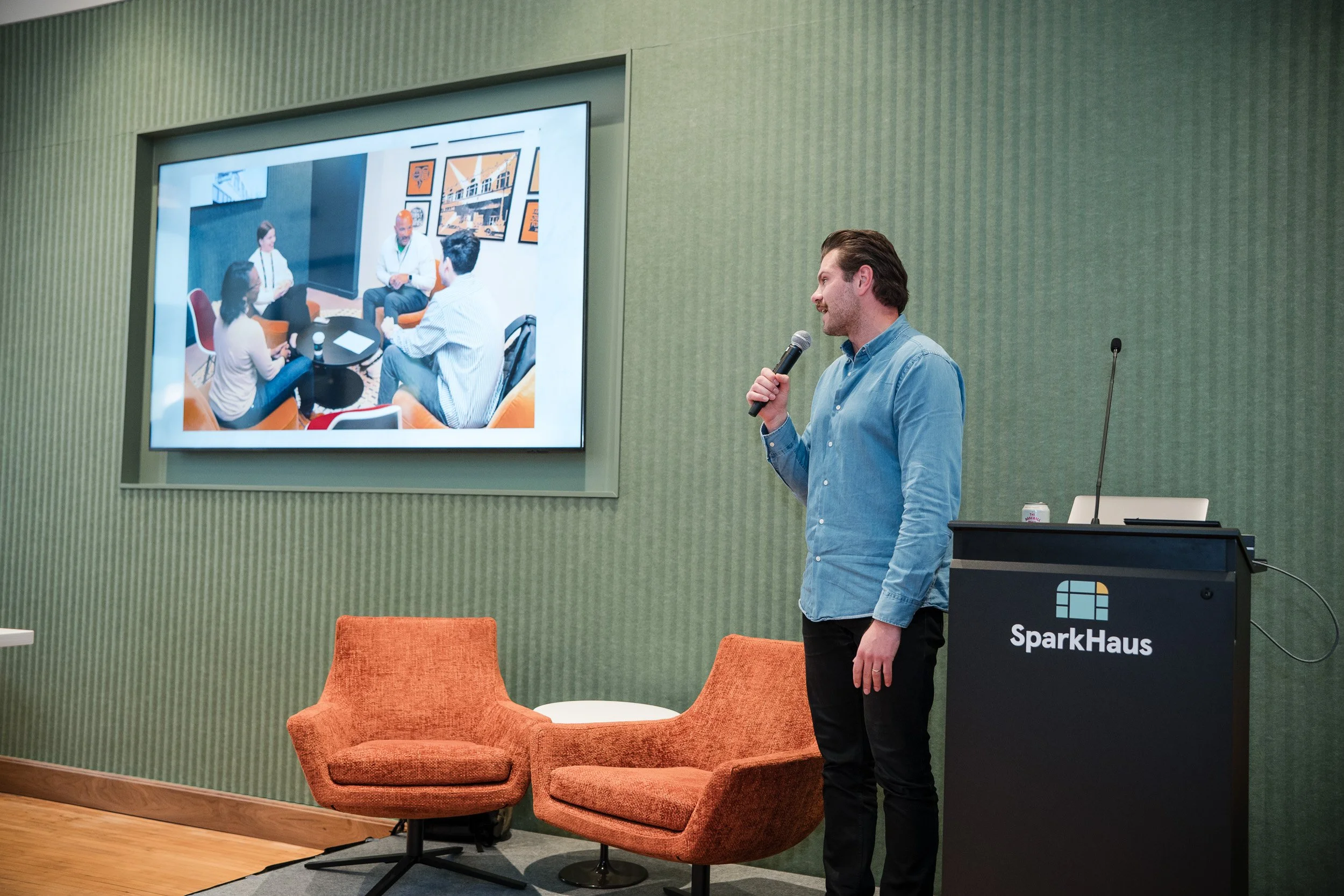 A man speaking into a microphone during a presentation at SparkHaus, with a large screen showing a photo of a group discussion. The room has green walls, orange chairs, and a podium.
