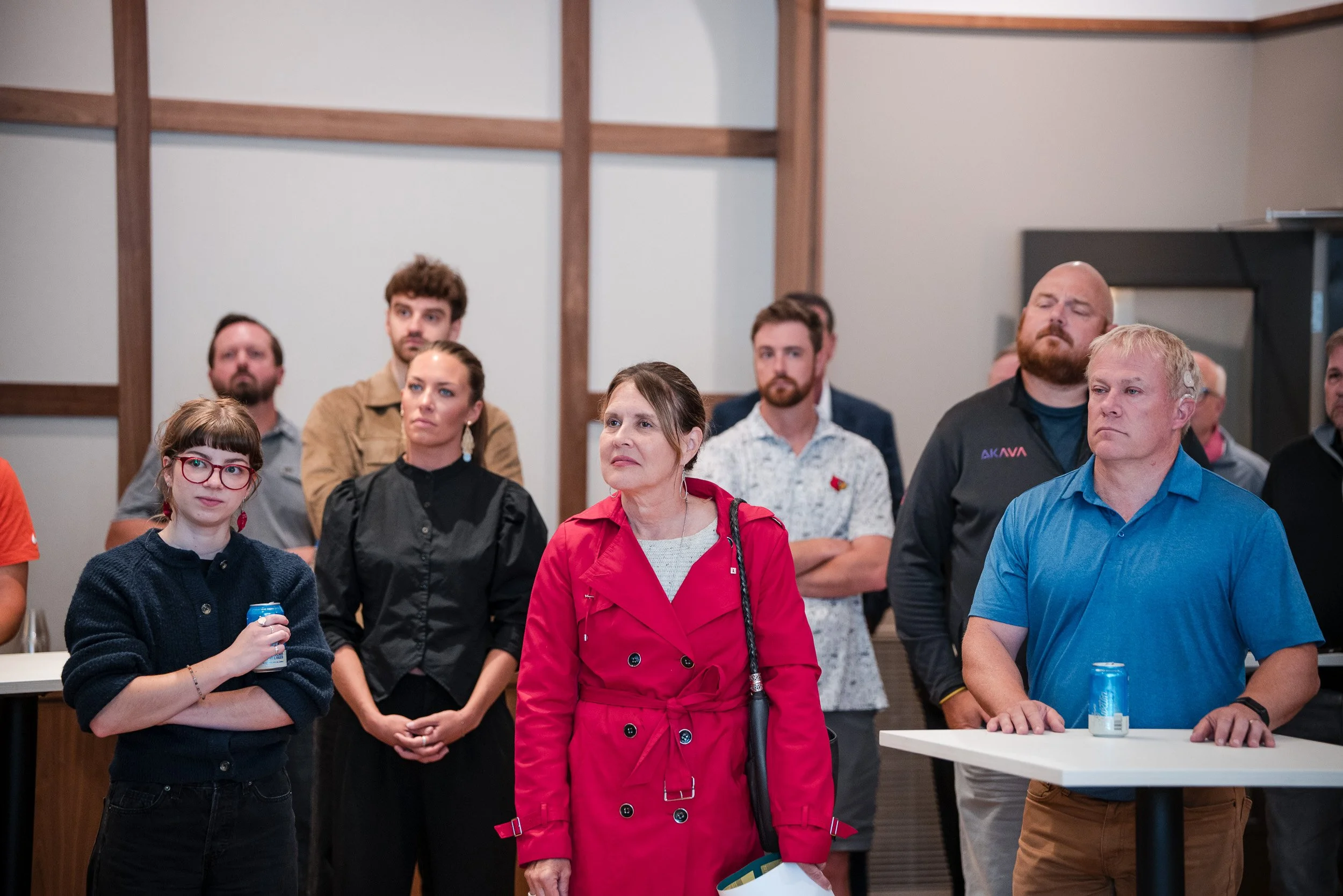 Group of adults and a girl attending an indoor event, standing and listening attentively, some with serious expressions.