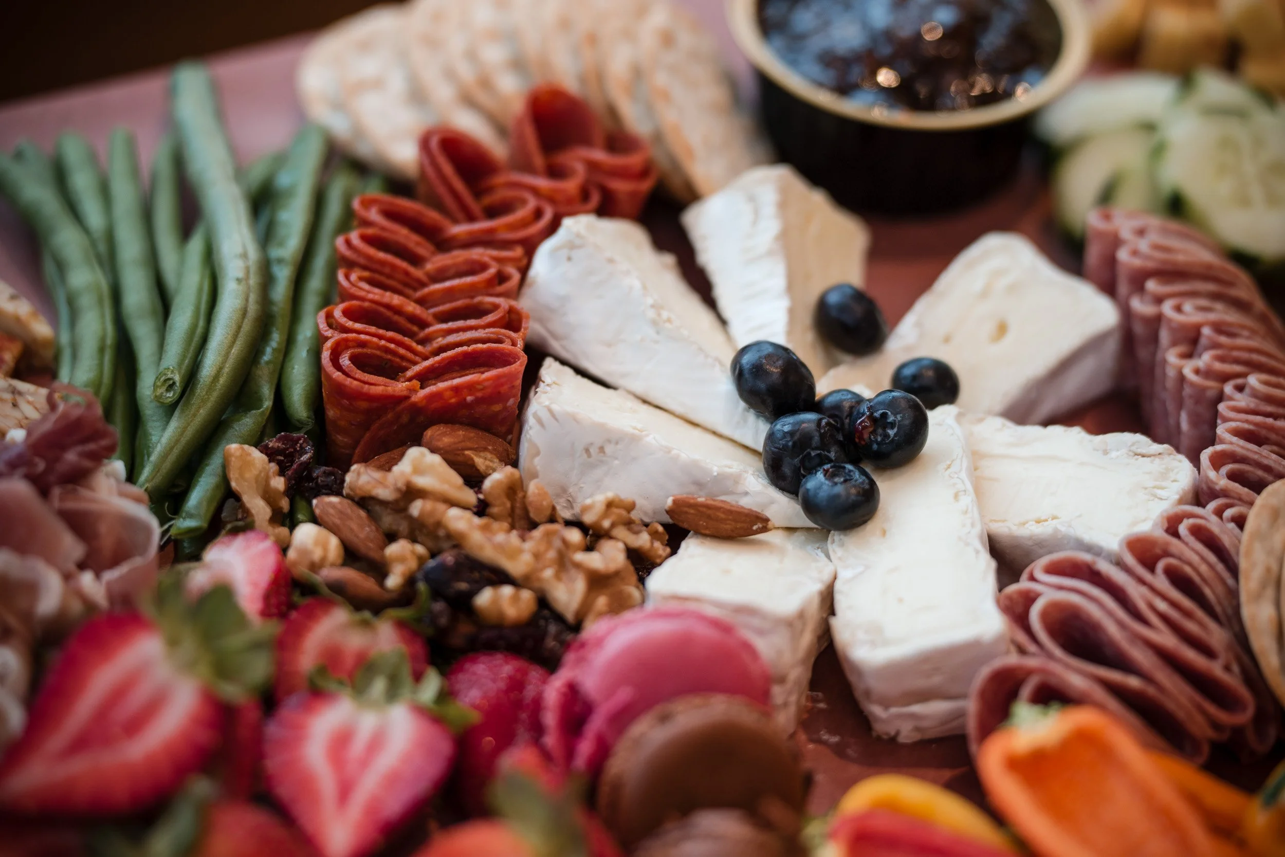 A cheese and charcuterie platter featuring sliced cheeses, rolled cured meats, fresh blueberries, strawberries, and various nuts, with a bowl of dark jam or sauce in the background.