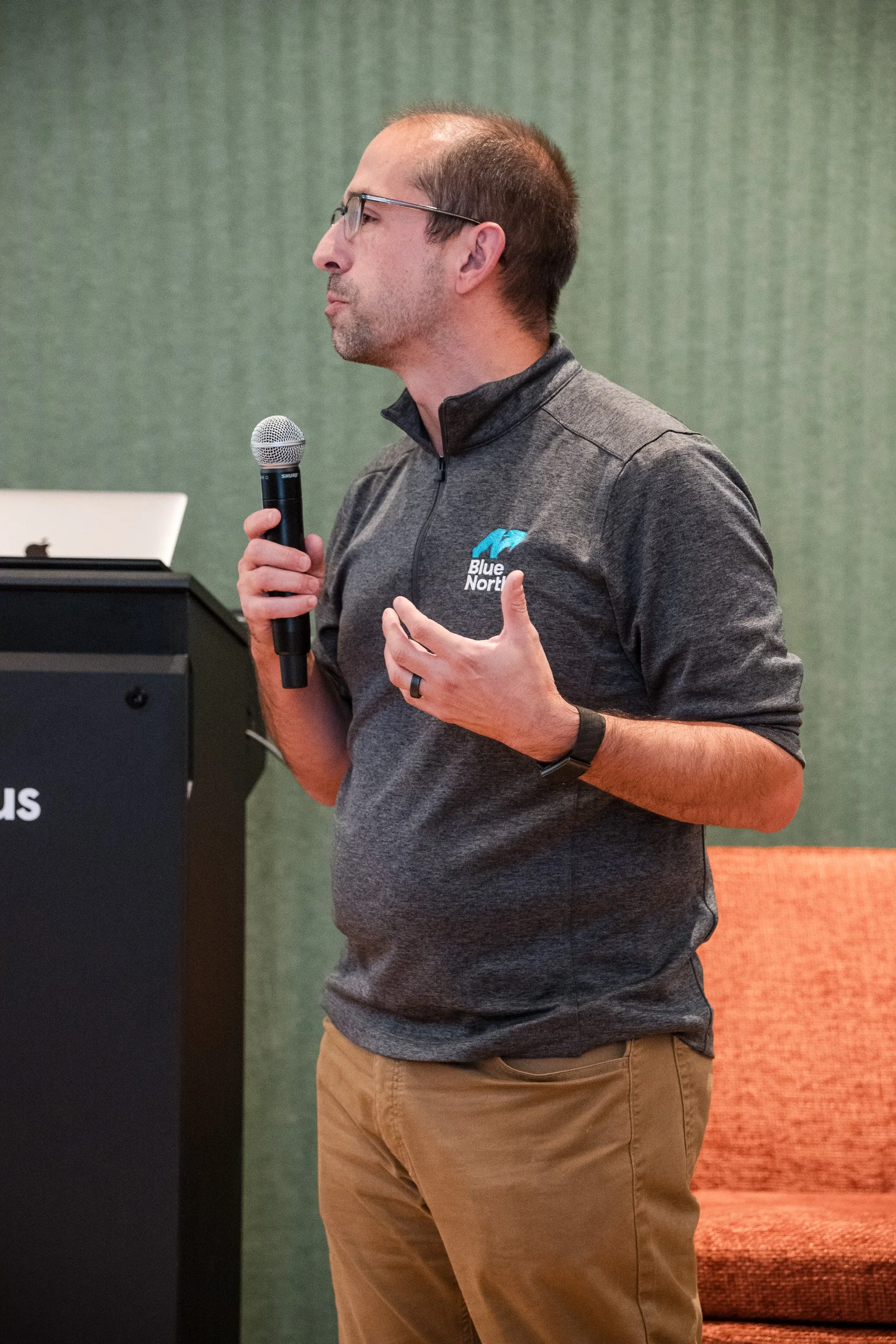 A man wearing glasses, a gray zip-up shirt with the Blue North logo, and khaki pants is speaking into a microphone while gesturing with his right hand in a presentation room with green walls and an orange chair.