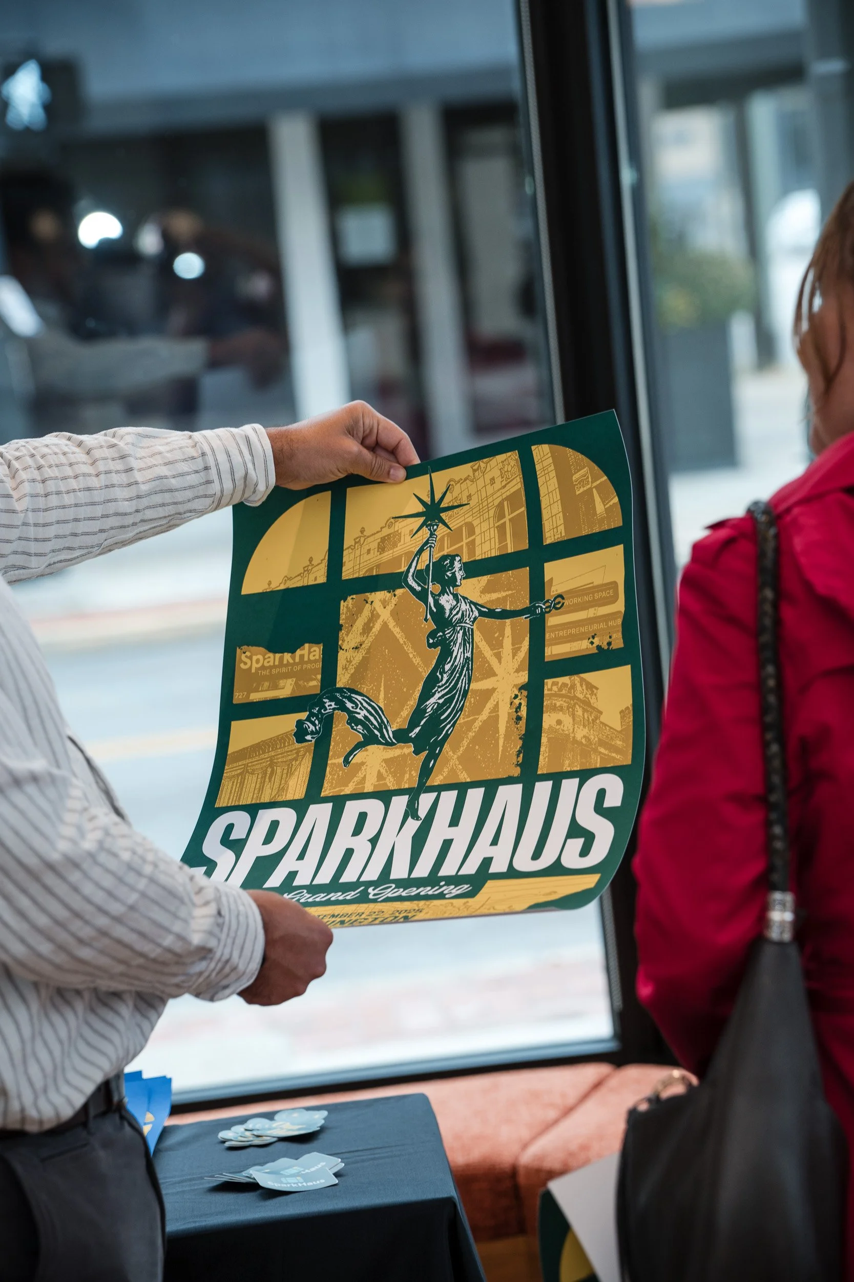 Person holding a green and yellow SparkHaus grand opening poster while another person in a red jacket looks on inside a building with large windows and a table with promotional materials.