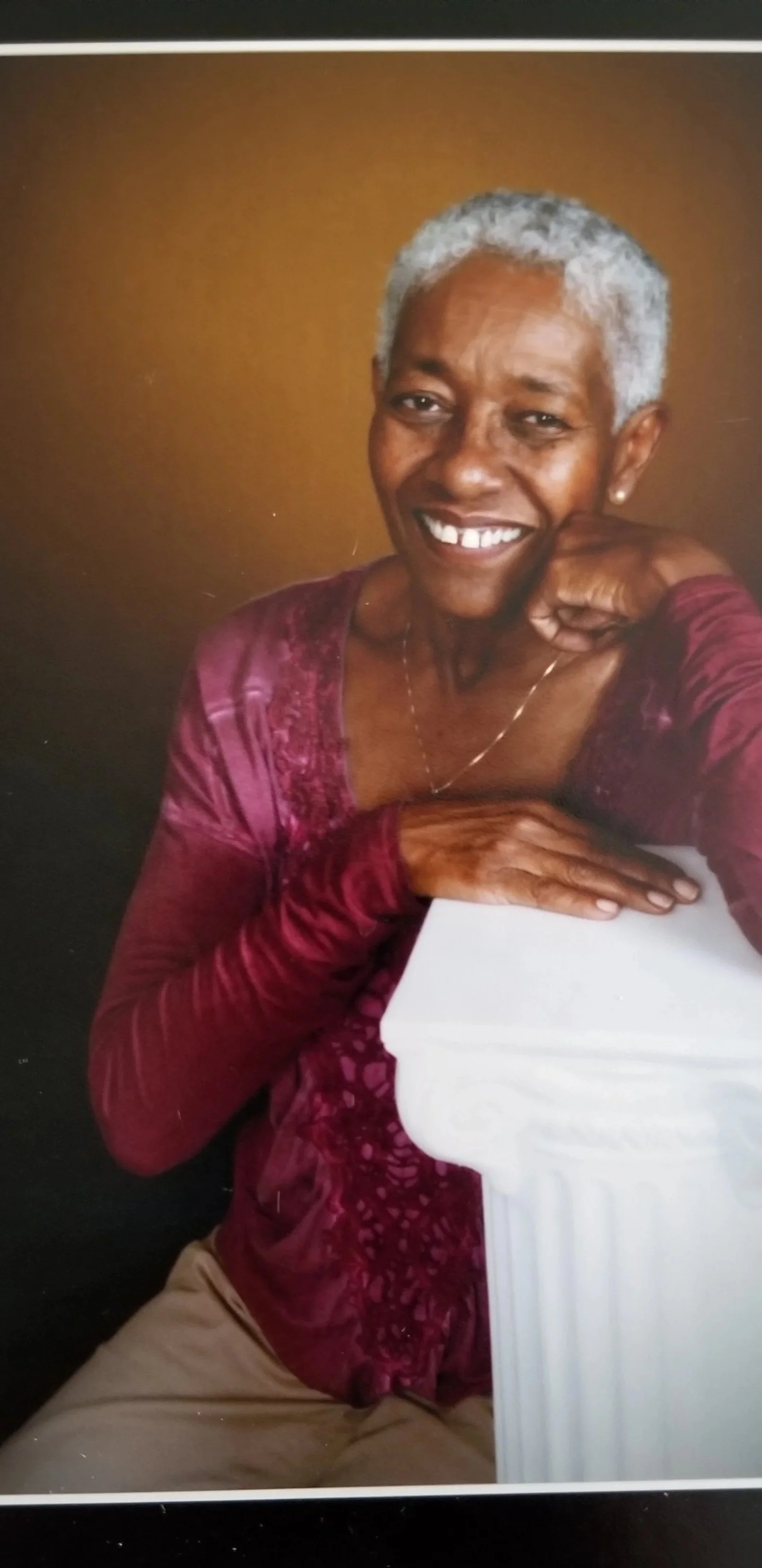 Portrait of an elderly woman with gray hair, smiling, leaning on a white column with a warm brown background.