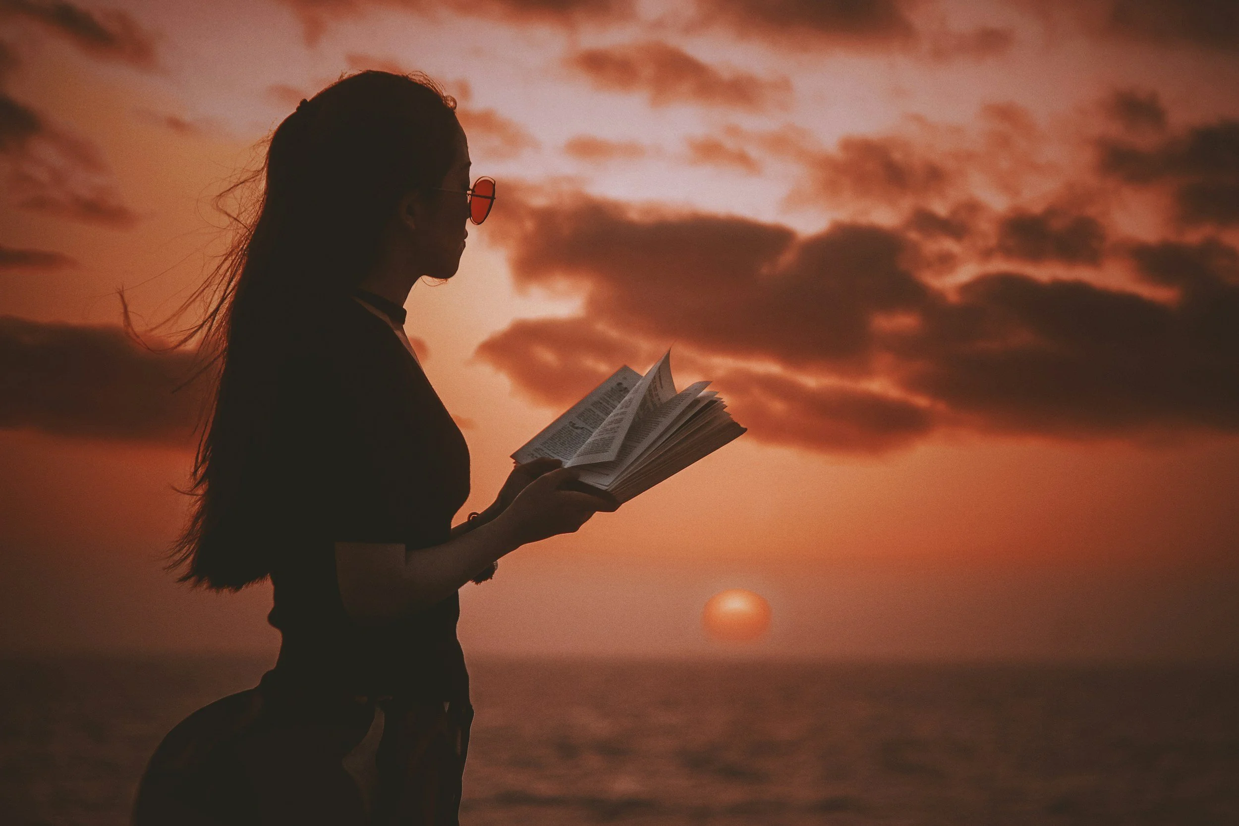 Silhouette of a woman reading a book outdoors during sunset or sunrise, with a cloudy sky and a glowing sun on the horizon.