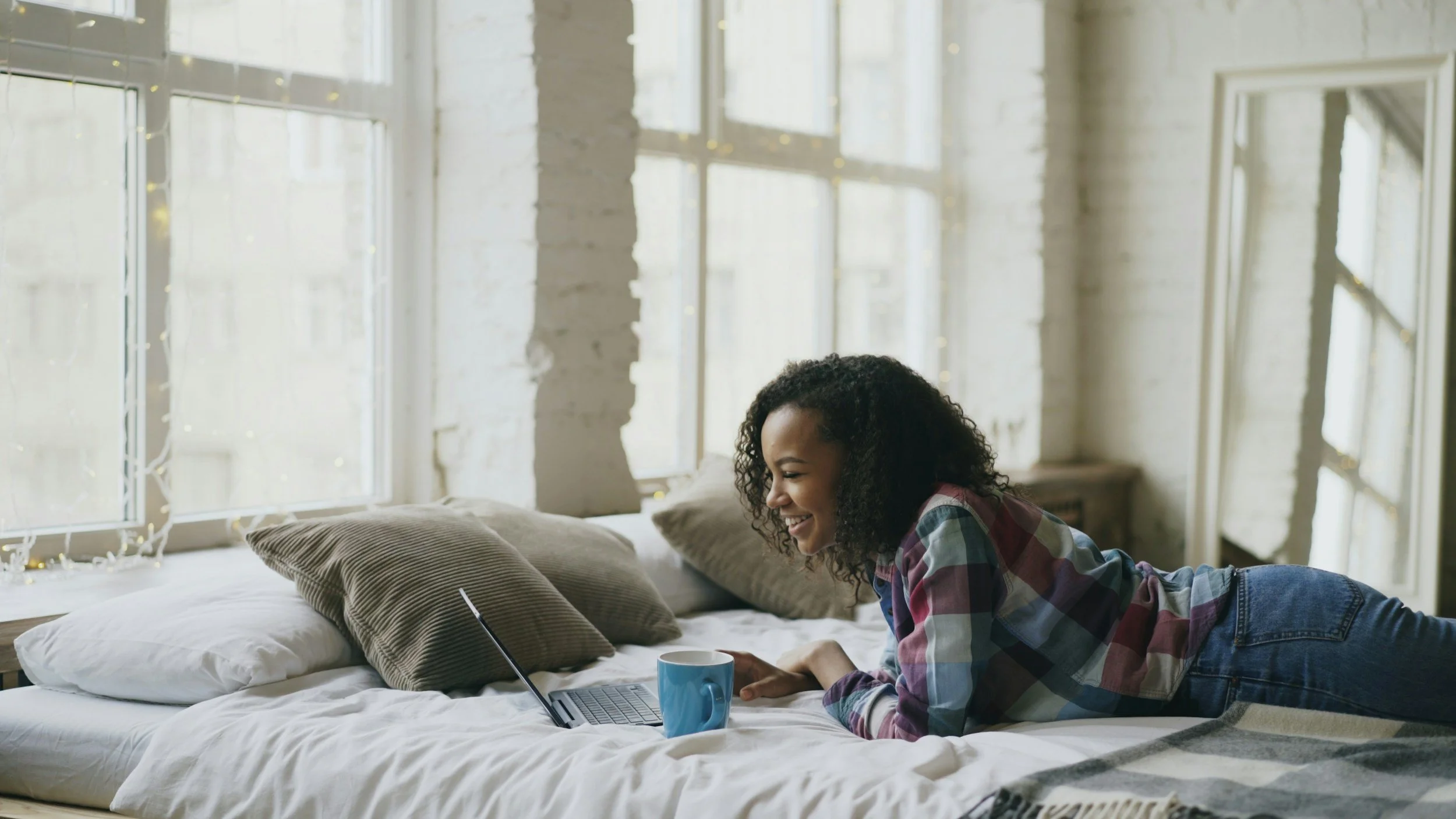 A young woman with curly hair lying on her stomach on a bed, smiling and looking at her laptop, with a ceramic mug nearby, in a bright room with large windows and white brick walls.