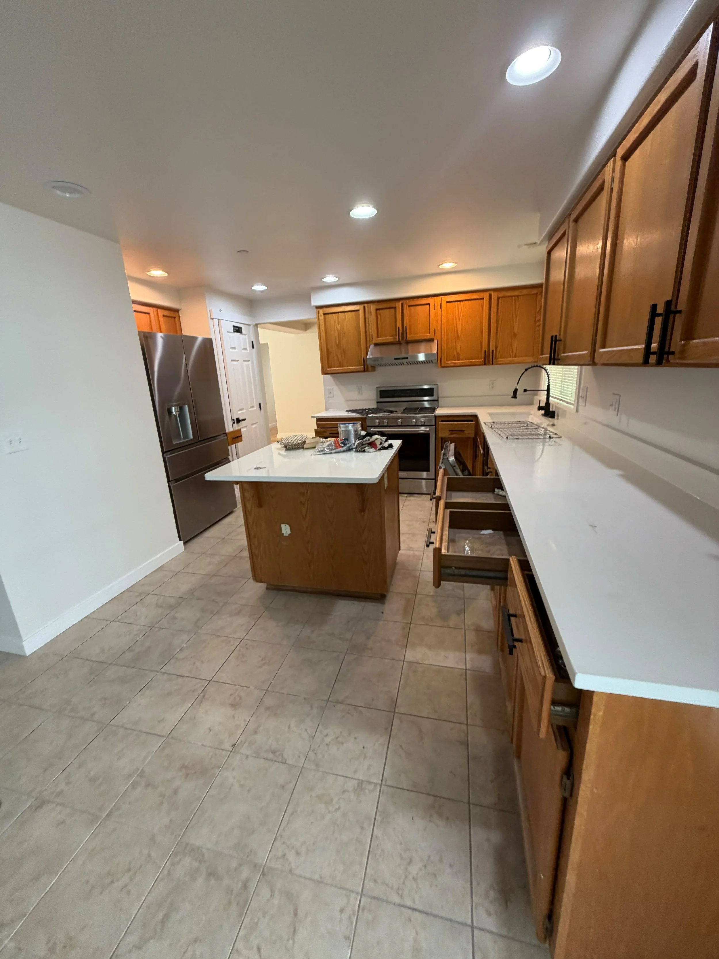 Kitchen with wooden cabinets, stainless steel refrigerator, white countertops, tiled floor, and a small window above the sink.