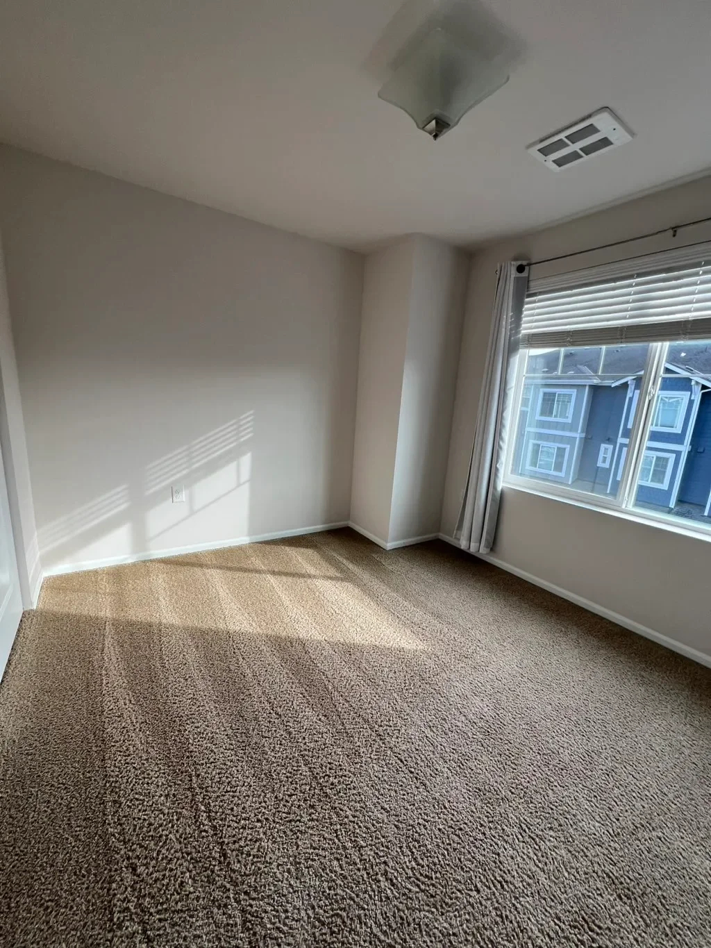 Empty bedroom with beige carpet, white walls, a large window with white blinds and curtains, and a ceiling light fixture.