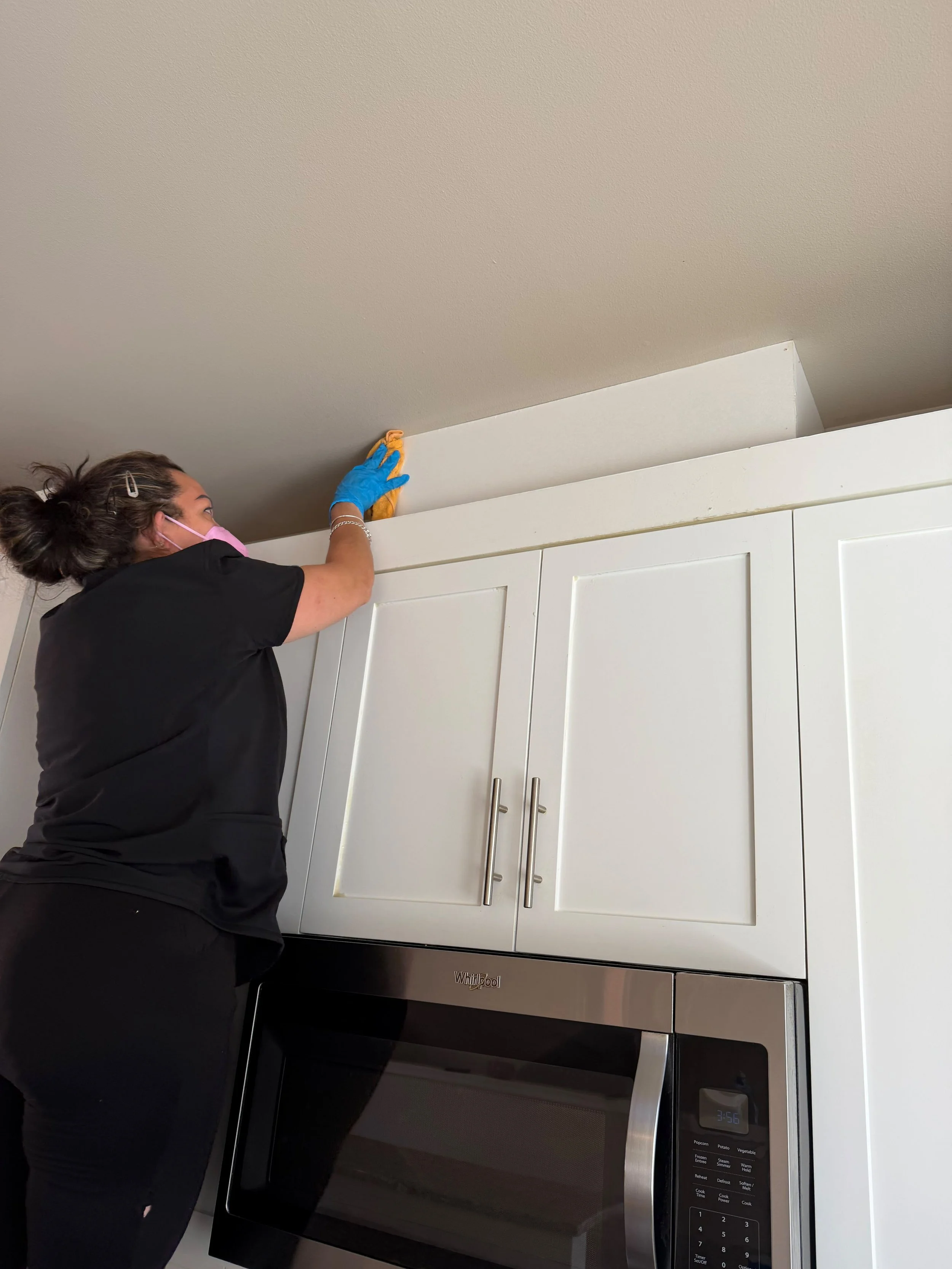 A woman wearing a pink face mask, blue gloves, and a hair clip, standing over a microwave oven in a kitchen, reaching toward the ceiling, possibly adjusting or installing something near the ceiling.