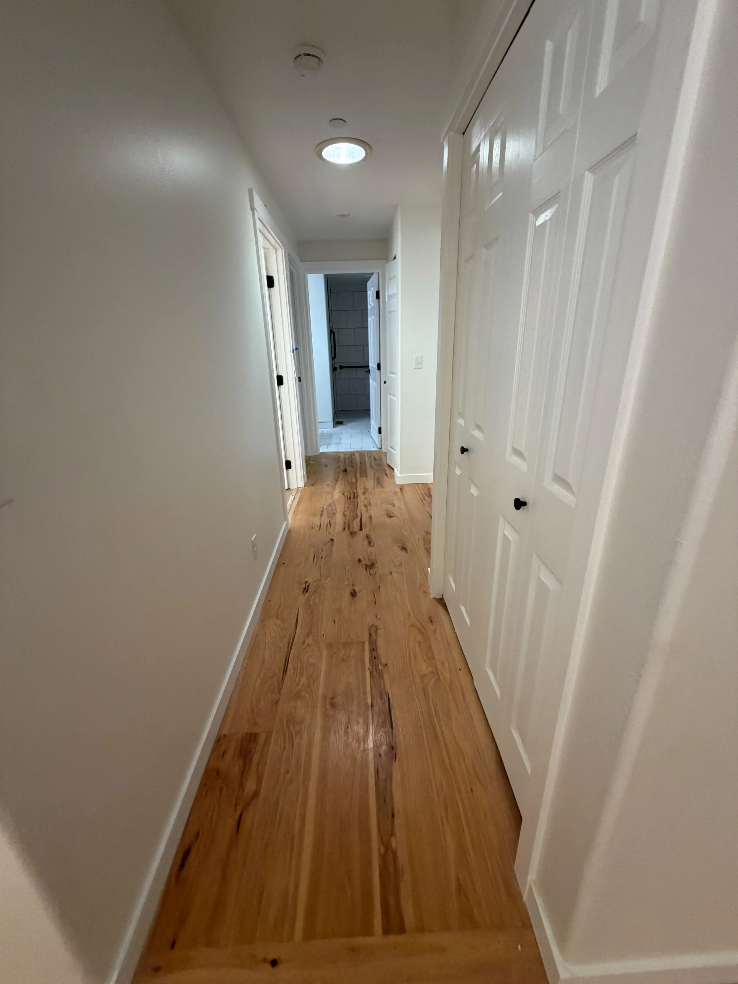 A hallway with white walls, wooden flooring, and a white door on the right side. There are ceiling lights and doors leading to different rooms, with a bathroom visible at the end of the hall.