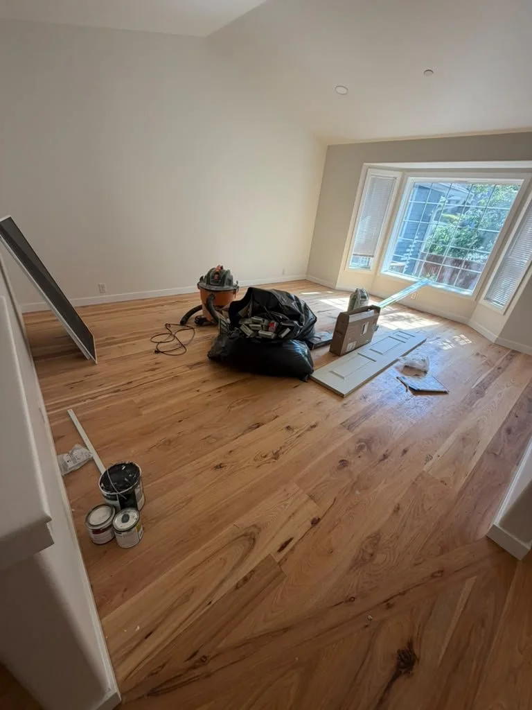 A room with hardwood flooring under construction, with tools, paint cans, and materials scattered around, and a large window letting in natural light.