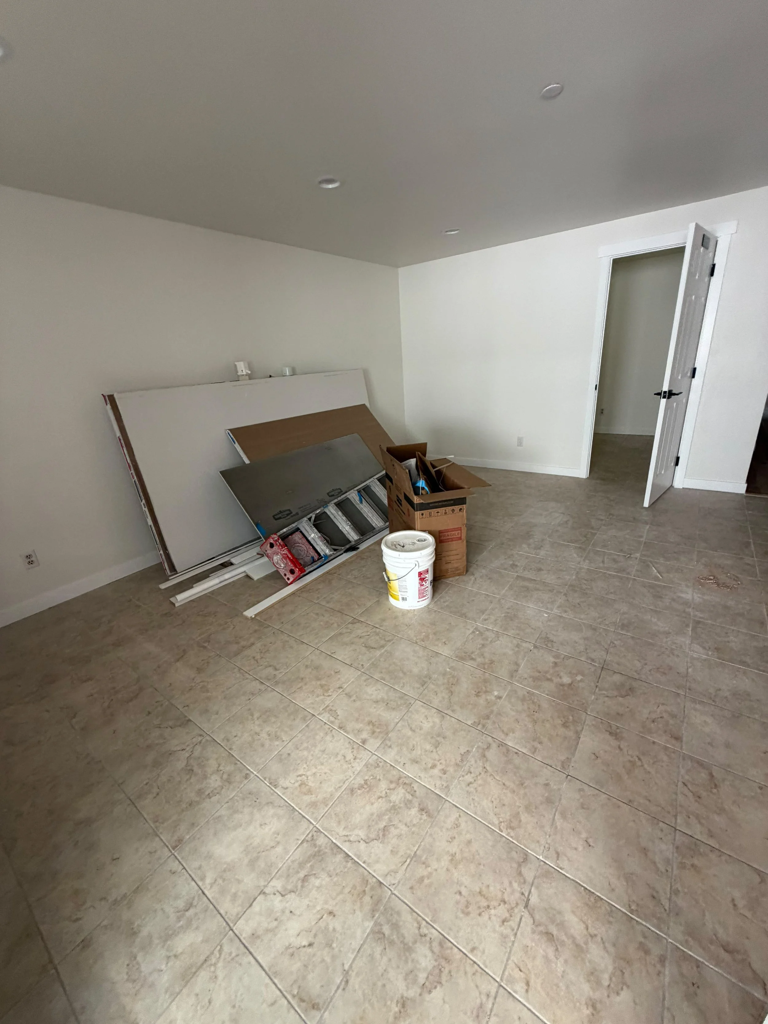 Empty room with beige tiled floor, white walls, a partially assembled bed frame leaning against the wall, and a cardboard box with packing materials on the floor.