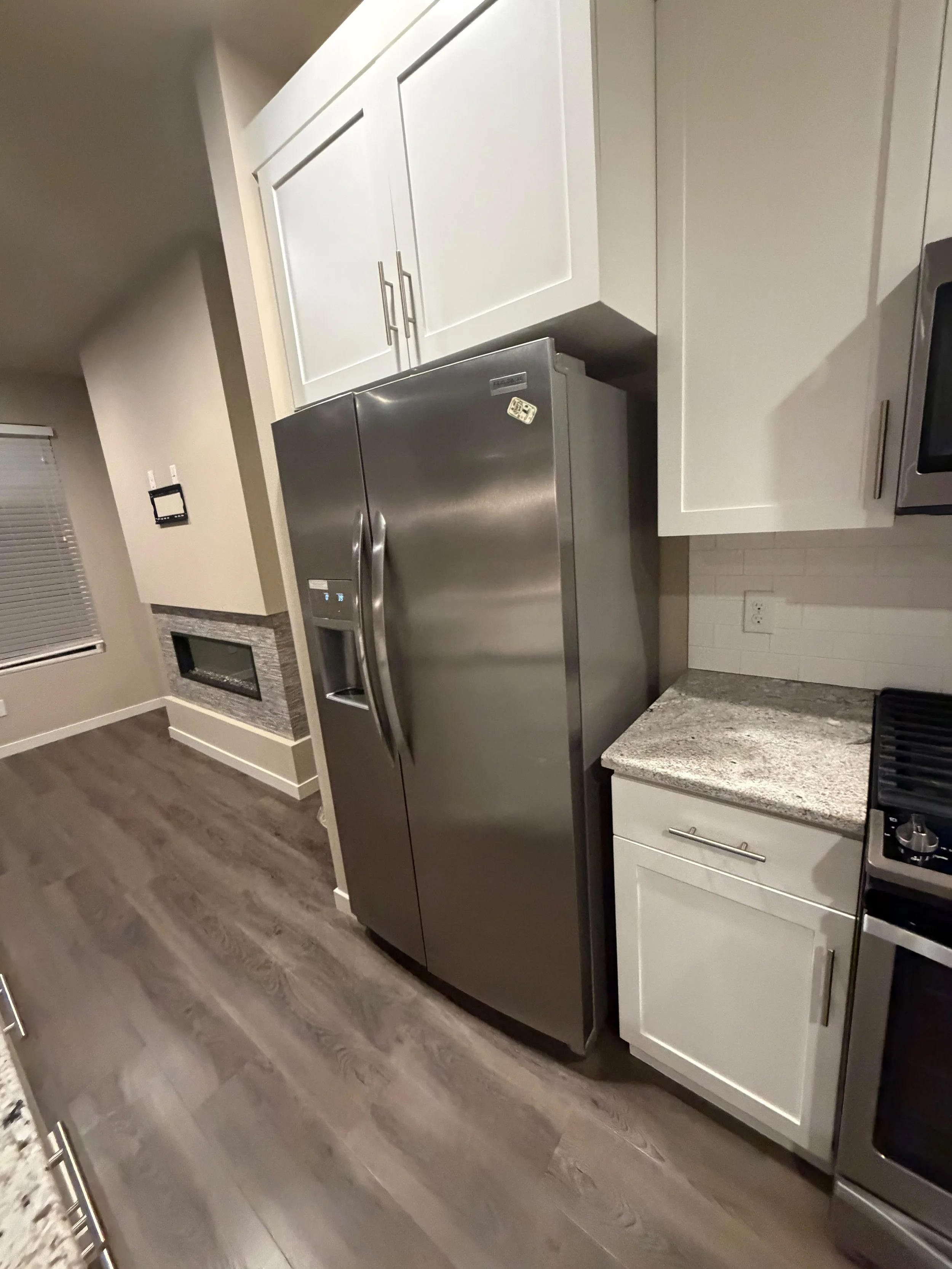 Modern kitchen with white cabinets, stainless steel refrigerator, granite countertop, and dark wood flooring.