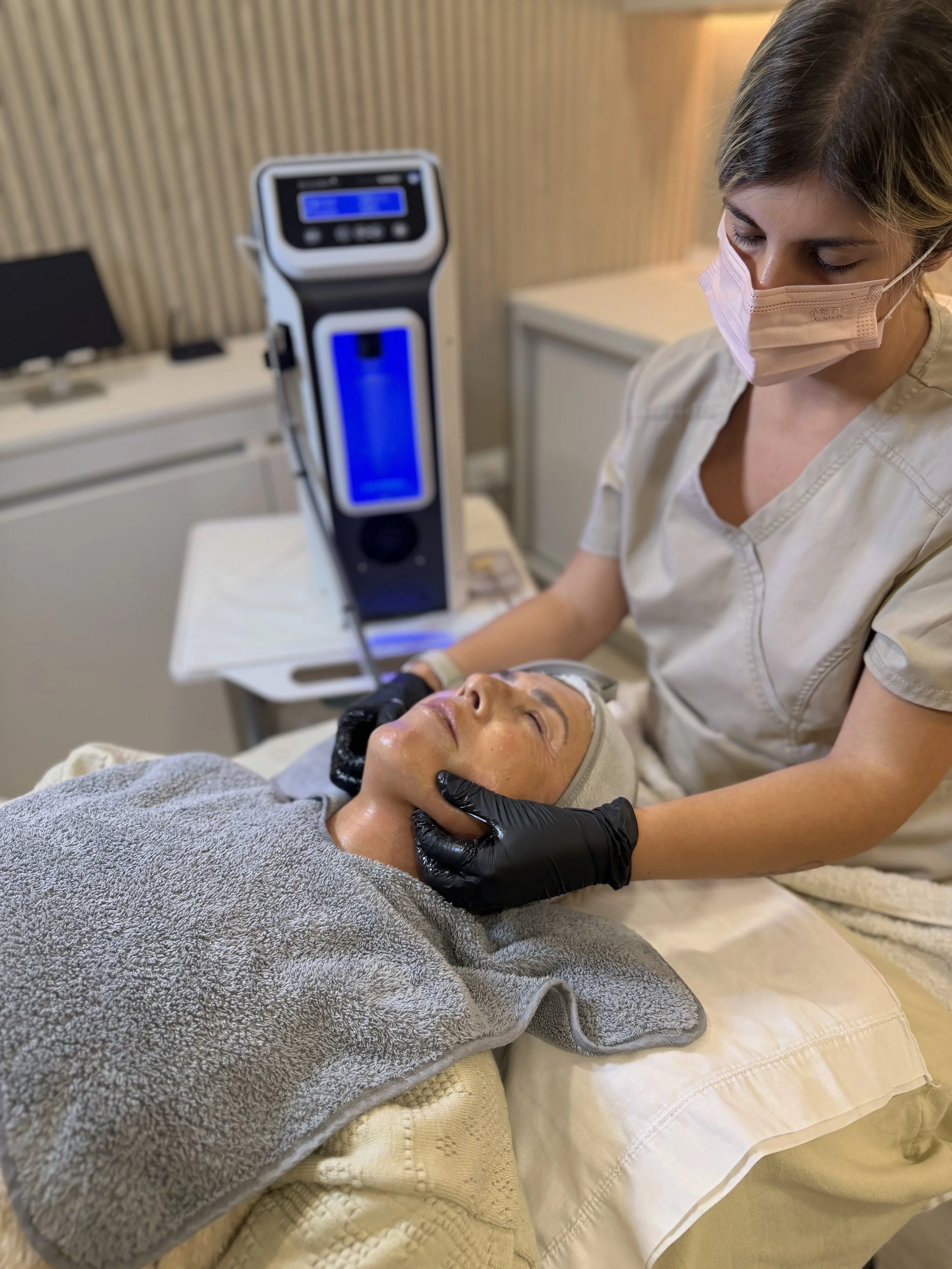 Una estética que realiza un tratamiento facial en una mujer acostada sobre la cama, con una toalla gris sobre el pecho y una manta. La mujer lleva una máscara facial y otra máscara en la cabeza. La esteticien usa guantes negros y un uniforme beige, y realiza una terapia mientras la mujer está tranquila con los ojos cerrados.