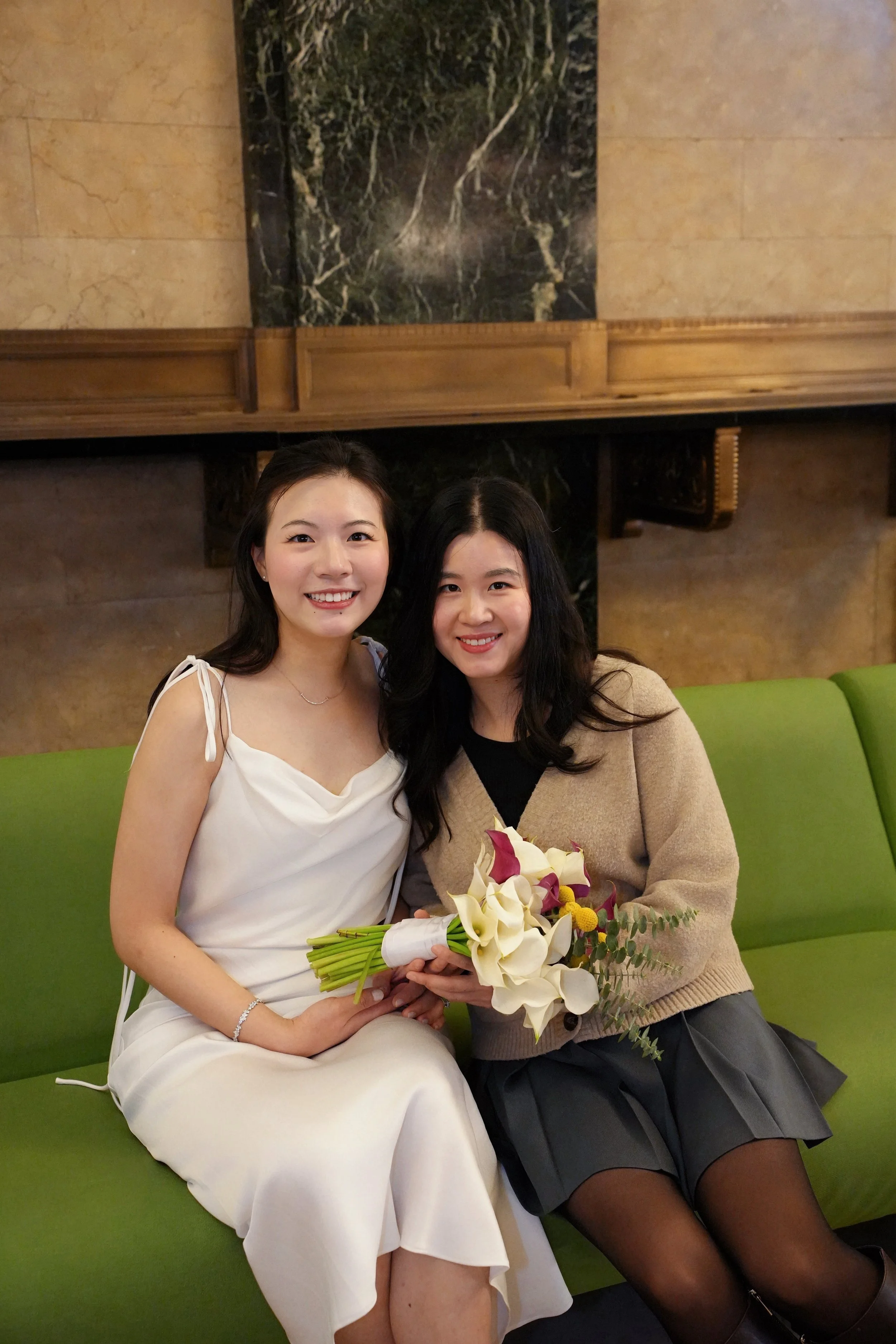 Two women sitting on a green bench, smiling. One in a white dress holding a bouquet of white and purple flowers, the other in a black skirt, beige sweater, and black stockings. They are in a room with wooden paneling and marble decor.