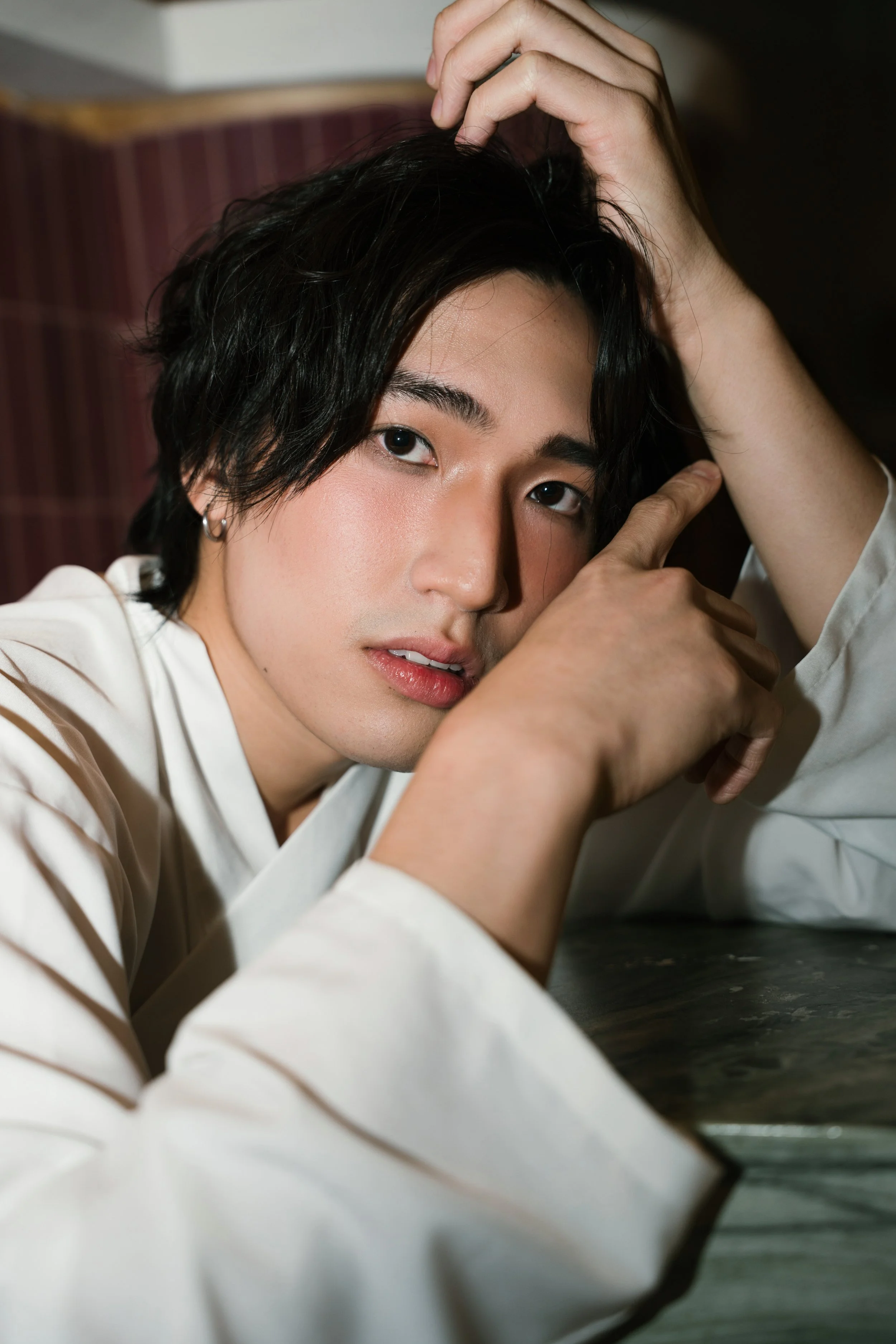 A young person with black hair, wearing earrings and a white shirt, is posing with their face close to the camera, hand touching their hair, appearing relaxed and contemplative.