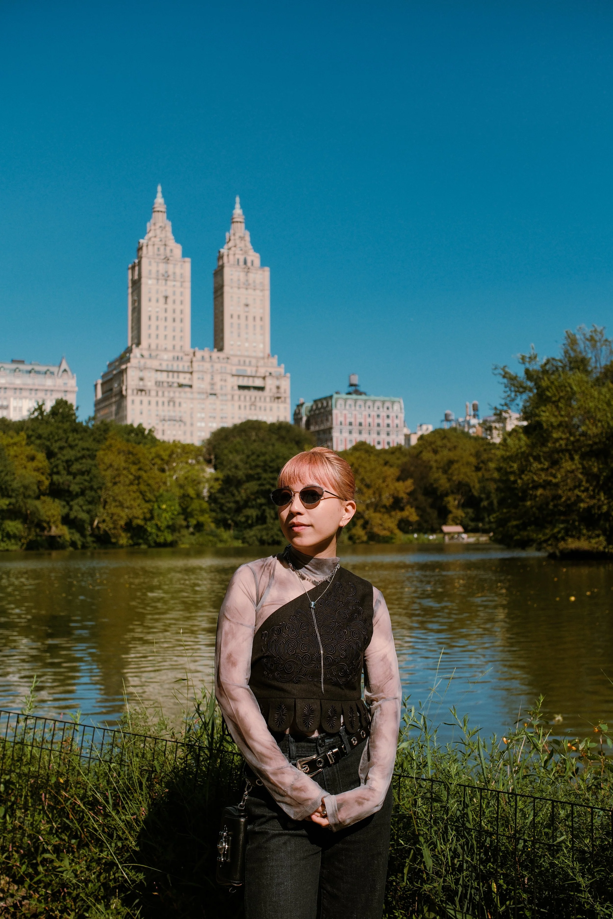 A young woman with short red hair and sunglasses standing by a lake with trees in the background. Tall buildings are visible in the distance against a clear blue sky.