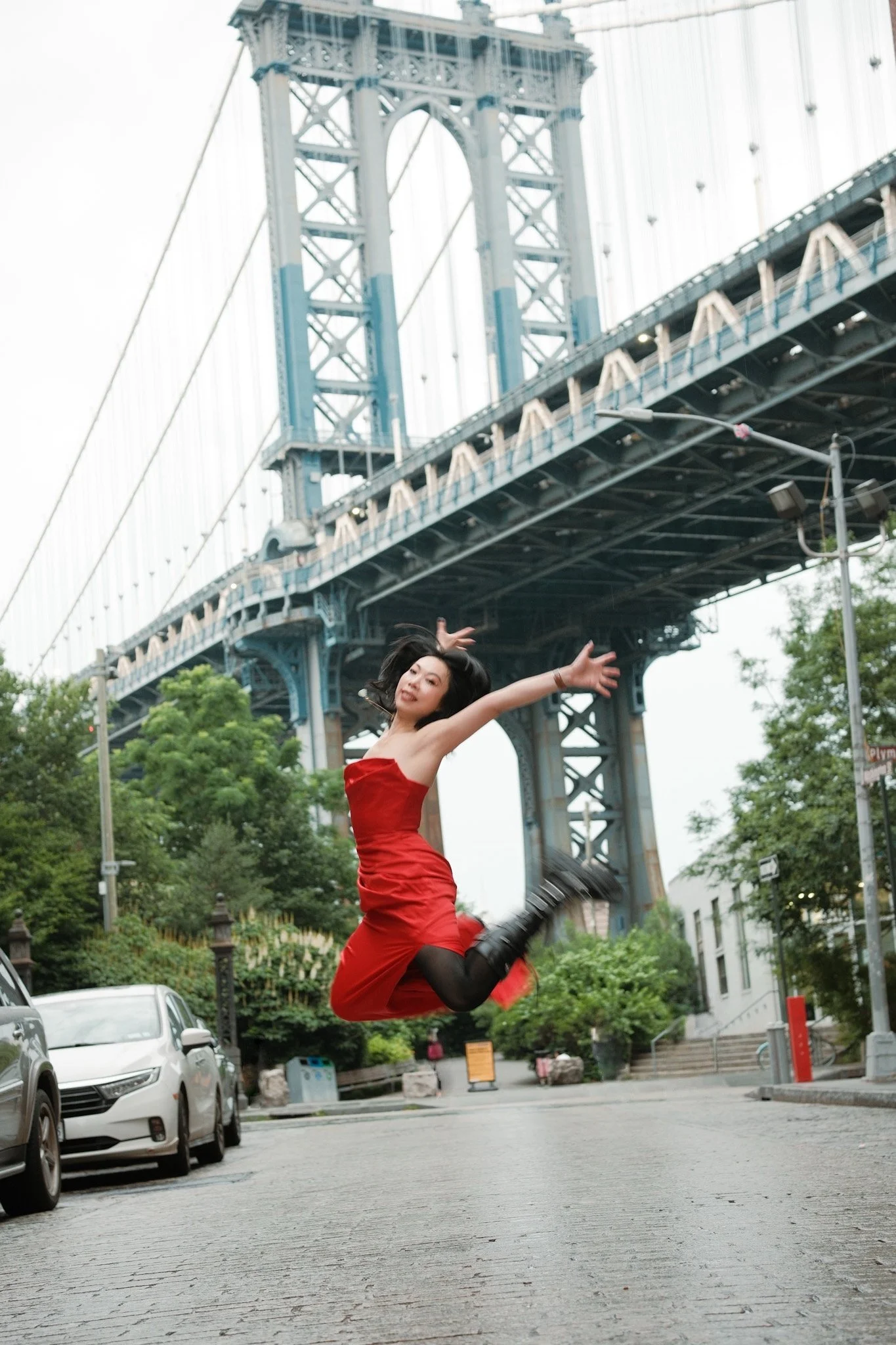 Woman in a red dress jumping with arms extended under a large bridge with suspension cables in an urban park setting.