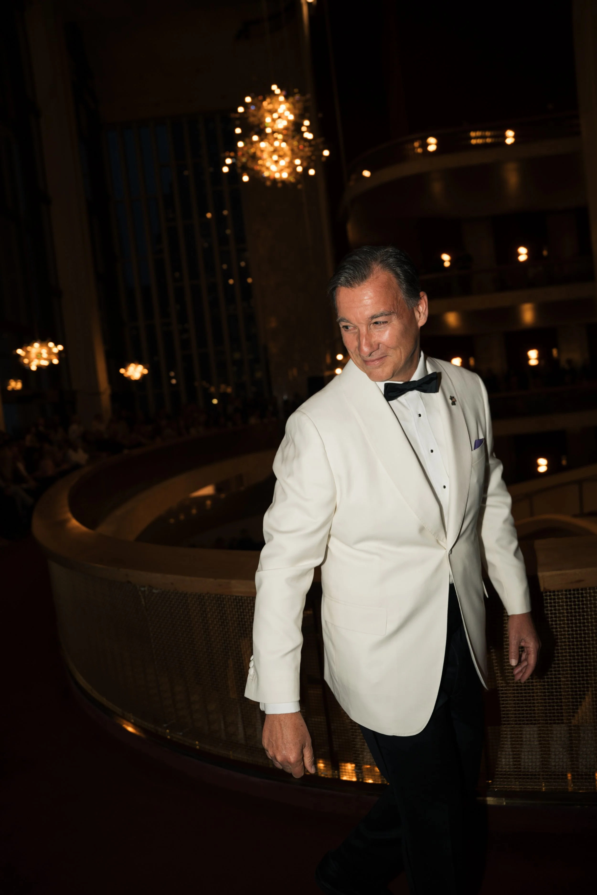 A man dressed in a white tuxedo jacket and black bow tie walking in a dimly lit venue with elegant chandeliers and a curved balcony in the background.