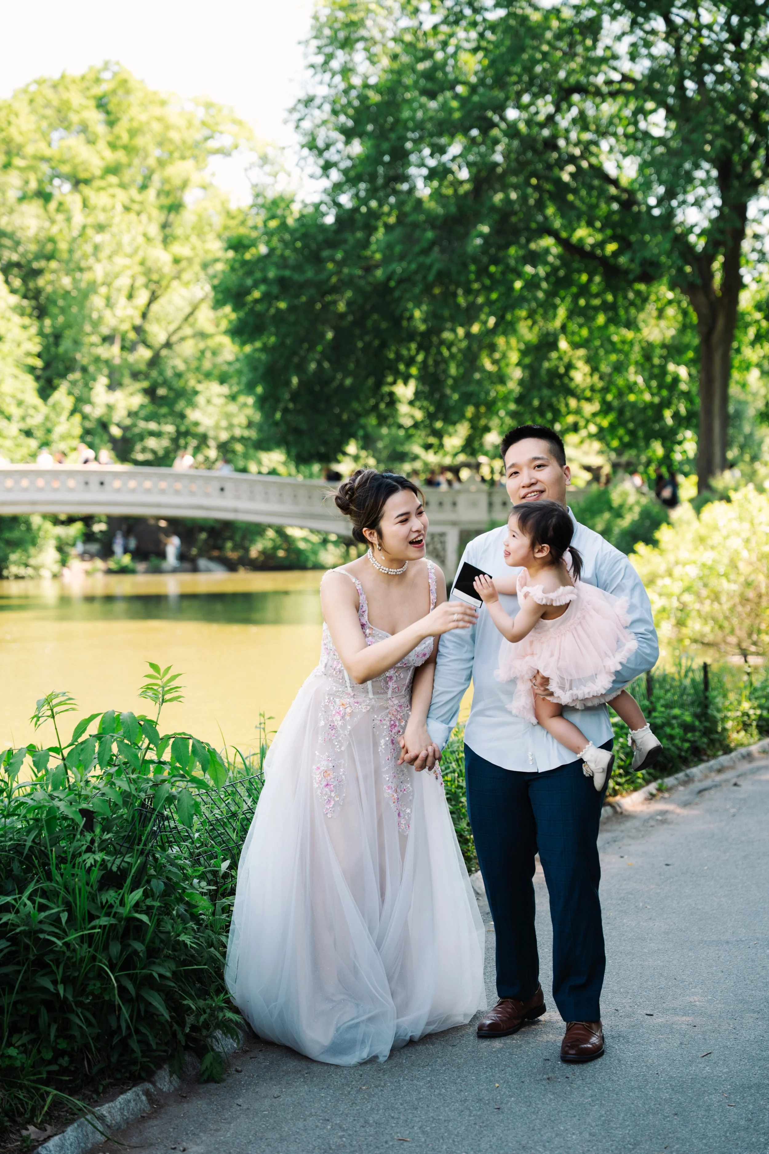 A family of three, including a woman in a wedding dress, a man, and a young girl, enjoying a walk in a park near a river with a stone bridge in the background. The trees are lush and green, indicating spring or summer.