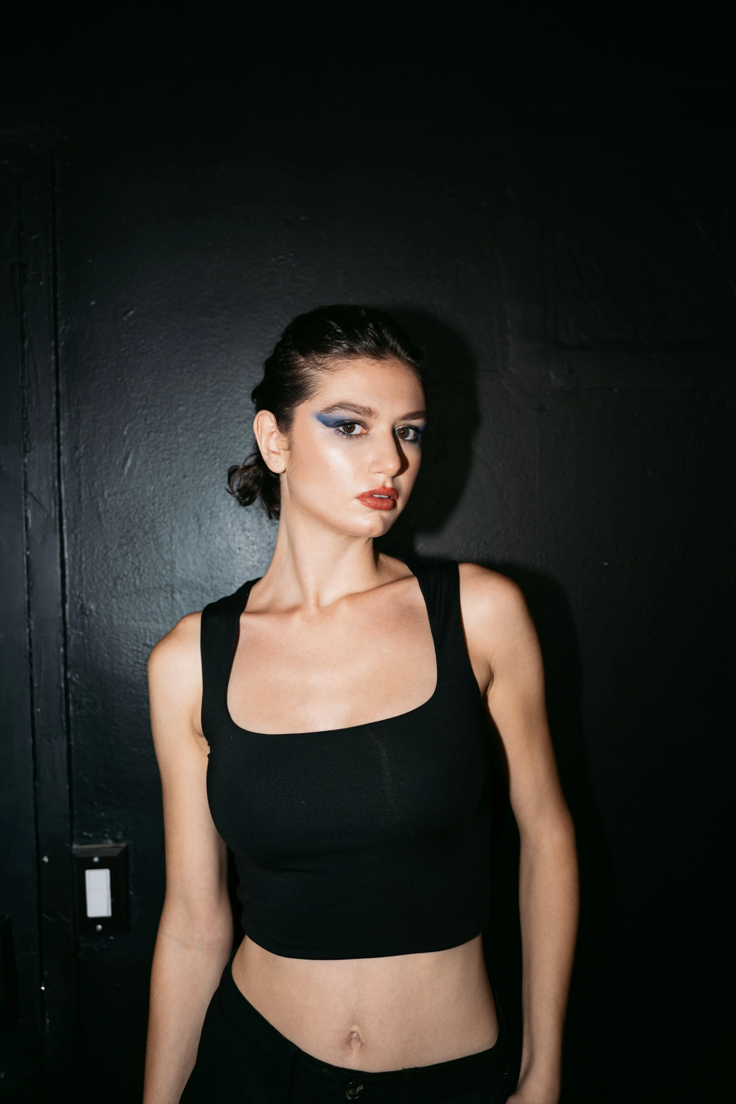 A young woman with dark hair styled in a low bun, wearing dramatic blue eye makeup, and a black sleeveless top, standing against a black wall with a serious expression.