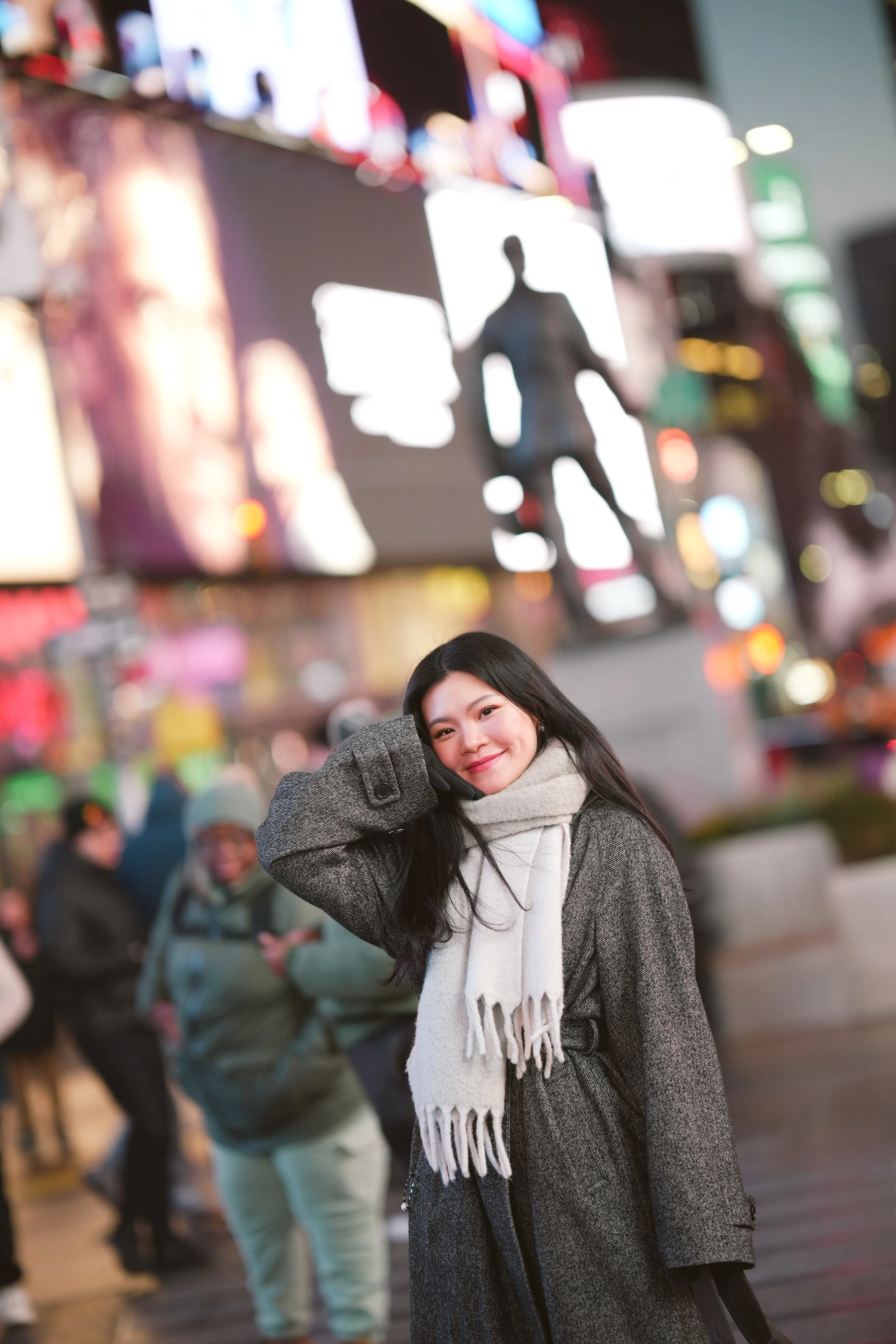 Smiling young woman with long dark hair wearing a gray coat and white scarf, posing in a busy city street at night with colorful blurred lights and billboards in the background.