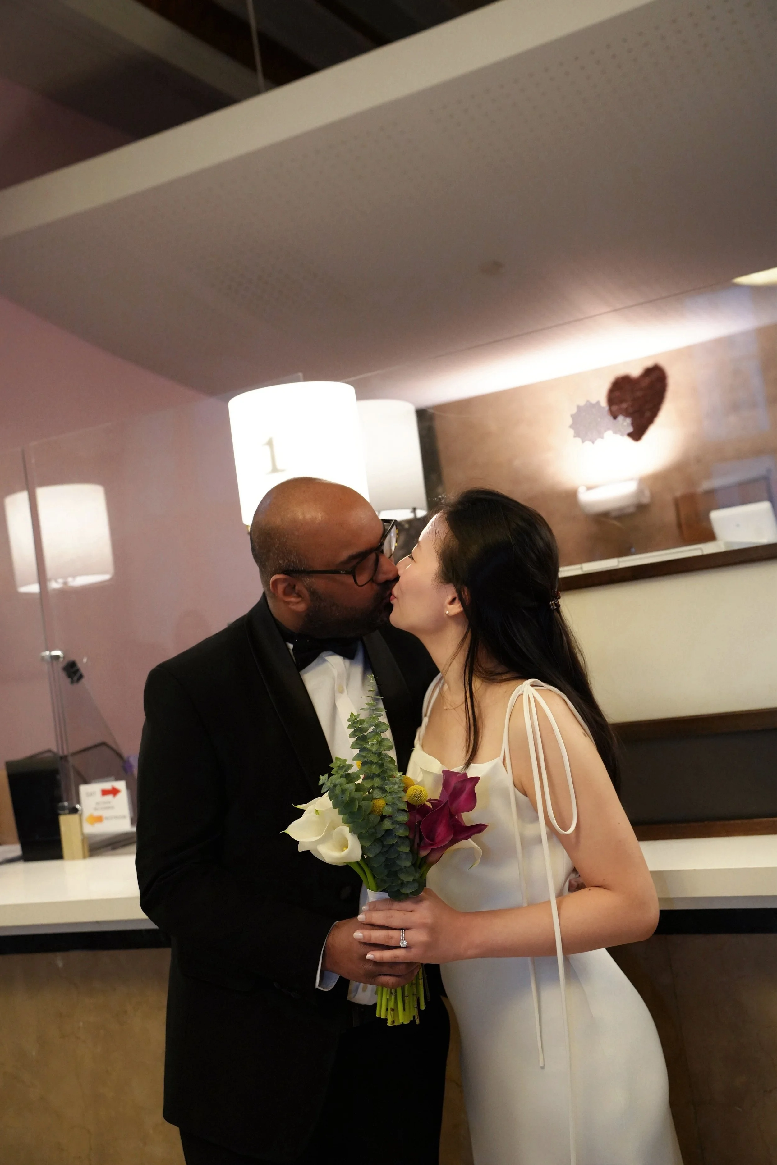 A couple in wedding attire kissing, the man in a black tuxedo and the woman in a white wedding gown holding a bouquet of flowers, in an indoor venue.