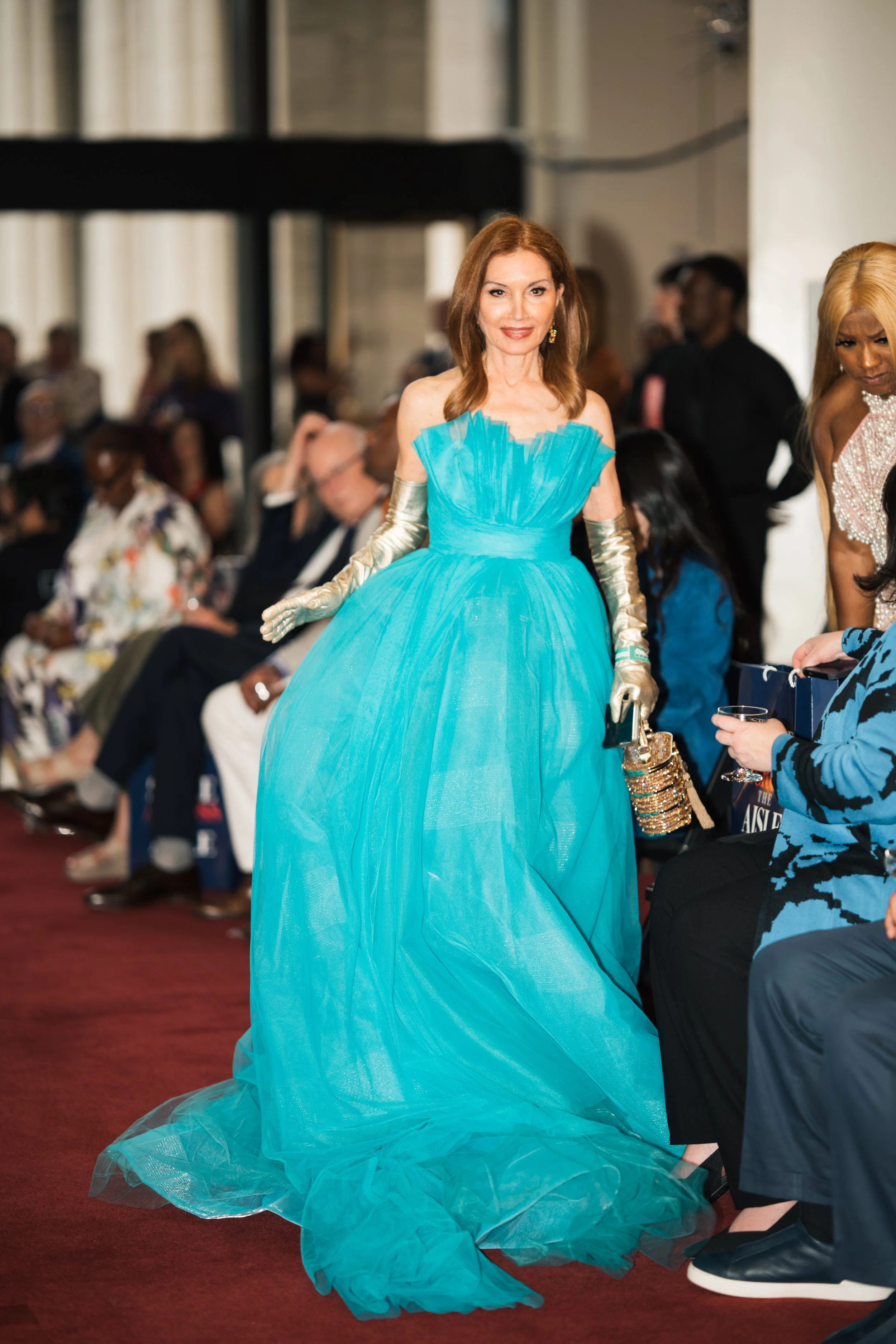 A woman in a bright blue, strapless, ball gown with tulle details at an event, standing and smiling, with a crowd seated in the background.