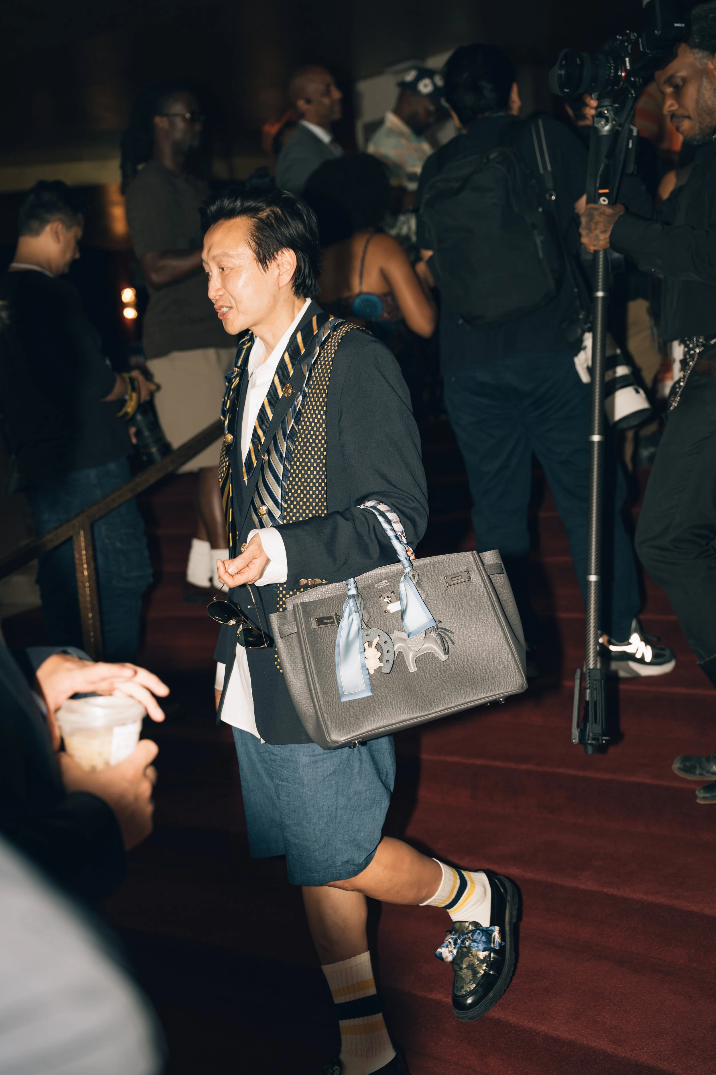 A man with dark hair wearing a gray blazer, white shirt, and blue shorts with striped knee-high socks, is walking through a crowded indoor event. He carries a large gray suitcase with a light blue ribbon, a black bag hanging from his arm, and sunglas