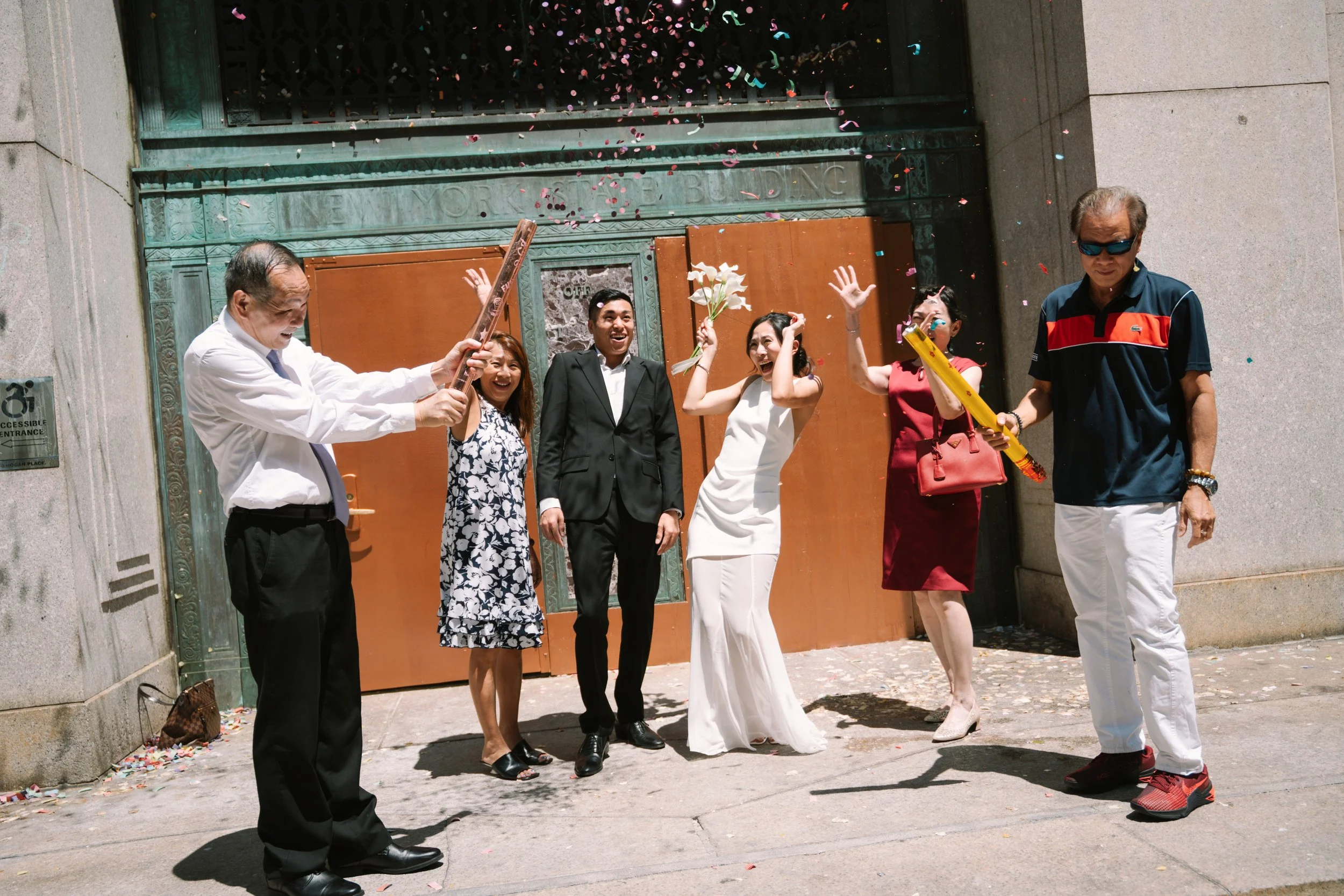 People celebrating outside a building with confetti and flowers, including a bride in a white dress, a groom in a black suit, and others dressed in colorful clothing.