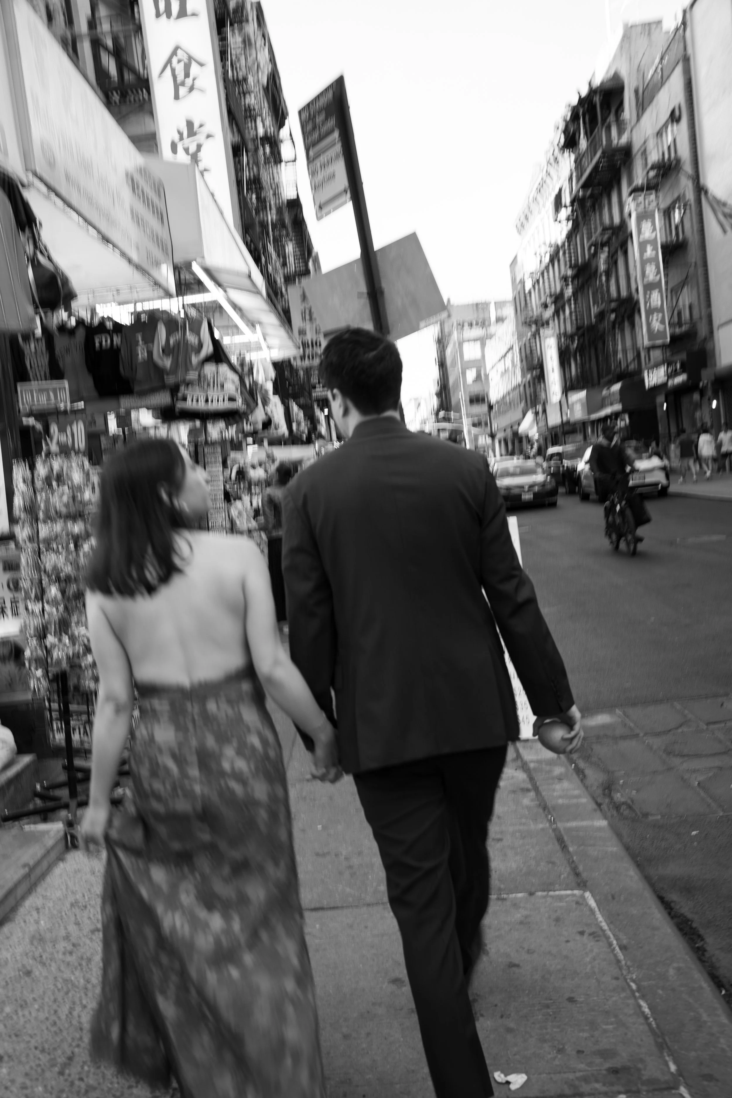 A black-and-white street scene with a man and woman holding hands walking down the sidewalk. The street is busy with cars, shops, and signs, with buildings lining both sides of the street.