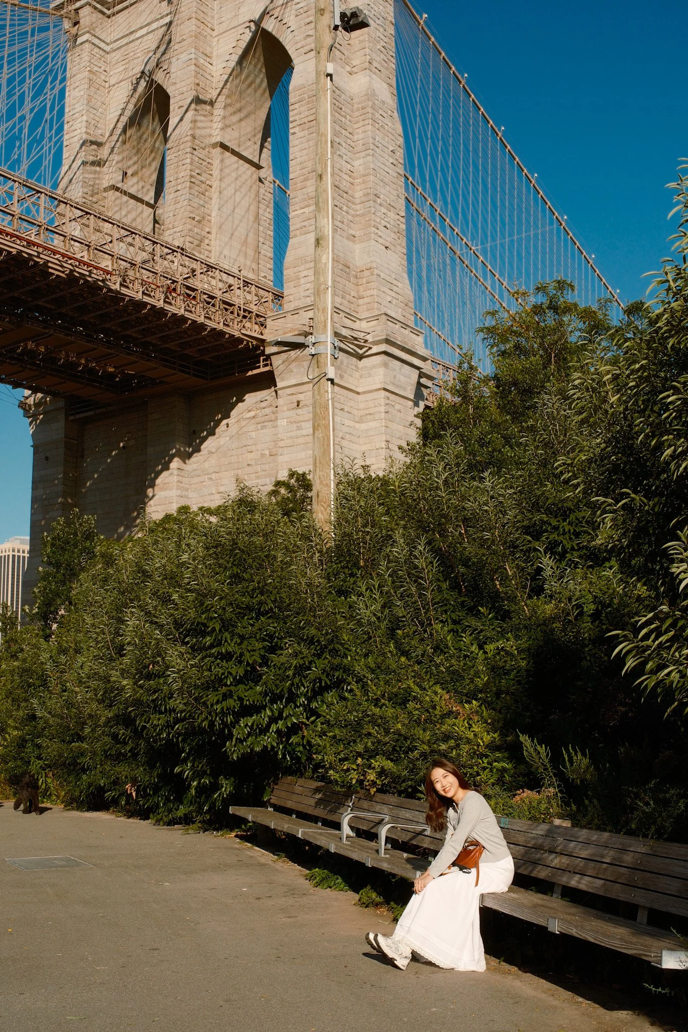 A woman with long brown hair, smiling, sitting on a wooden park bench in a park with green trees, near the Brooklyn Bridge in New York City.
