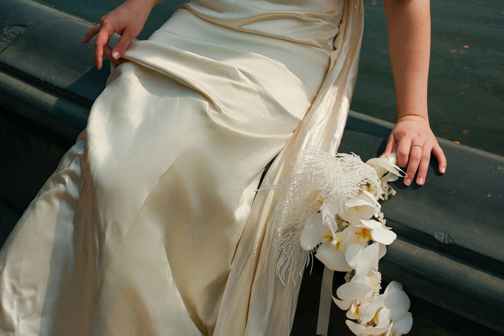 Close-up of a person wearing a silky, cream-colored dress, sitting on a dark bench near water, with a bouquet of white orchids and lace ribbons resting on the bench.