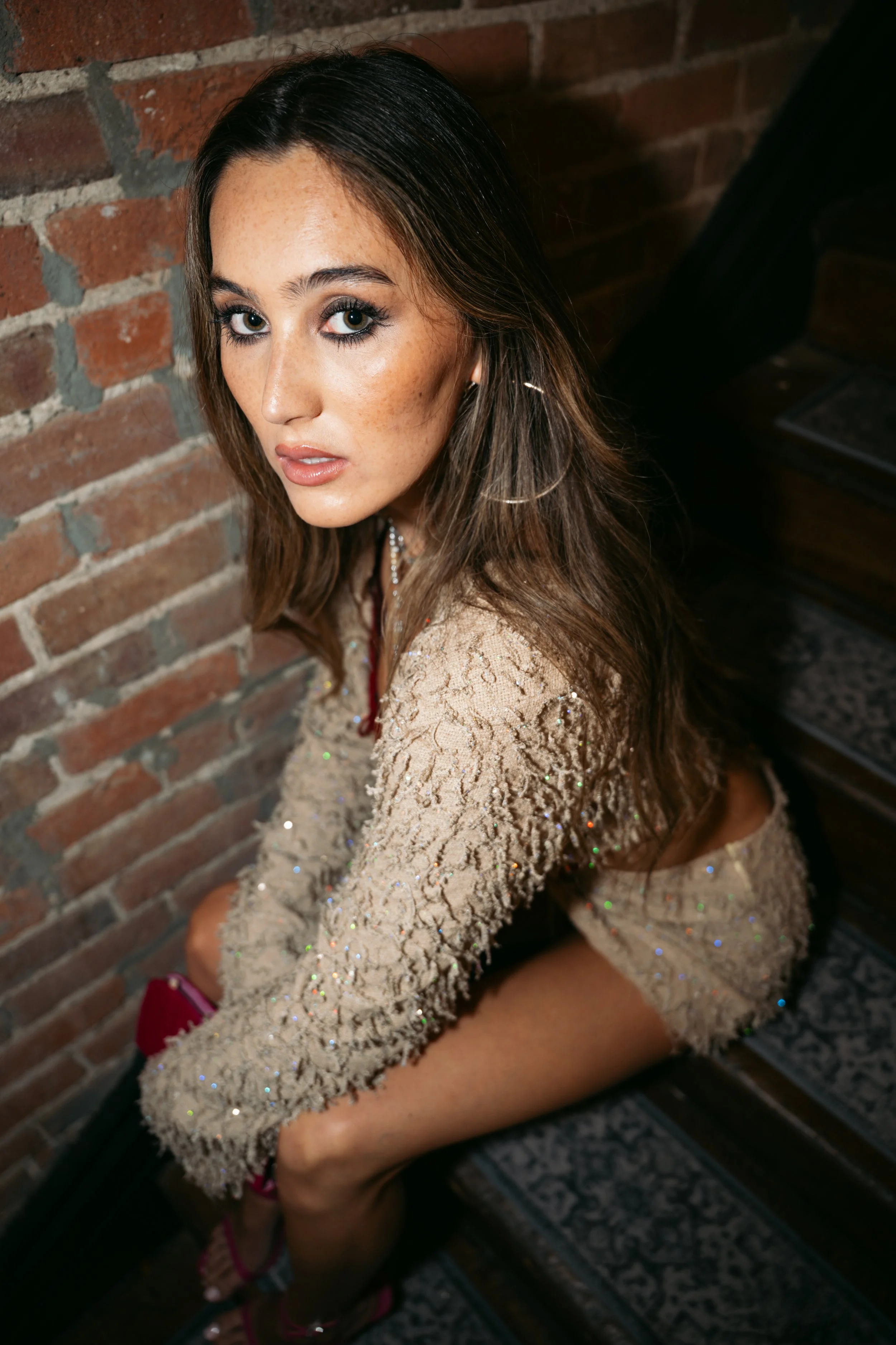 A young woman with long dark hair, light skin, and freckles, wearing makeup, a sparkly beige jacket, and pink shoes, sitting on a bench against an exposed brick wall.