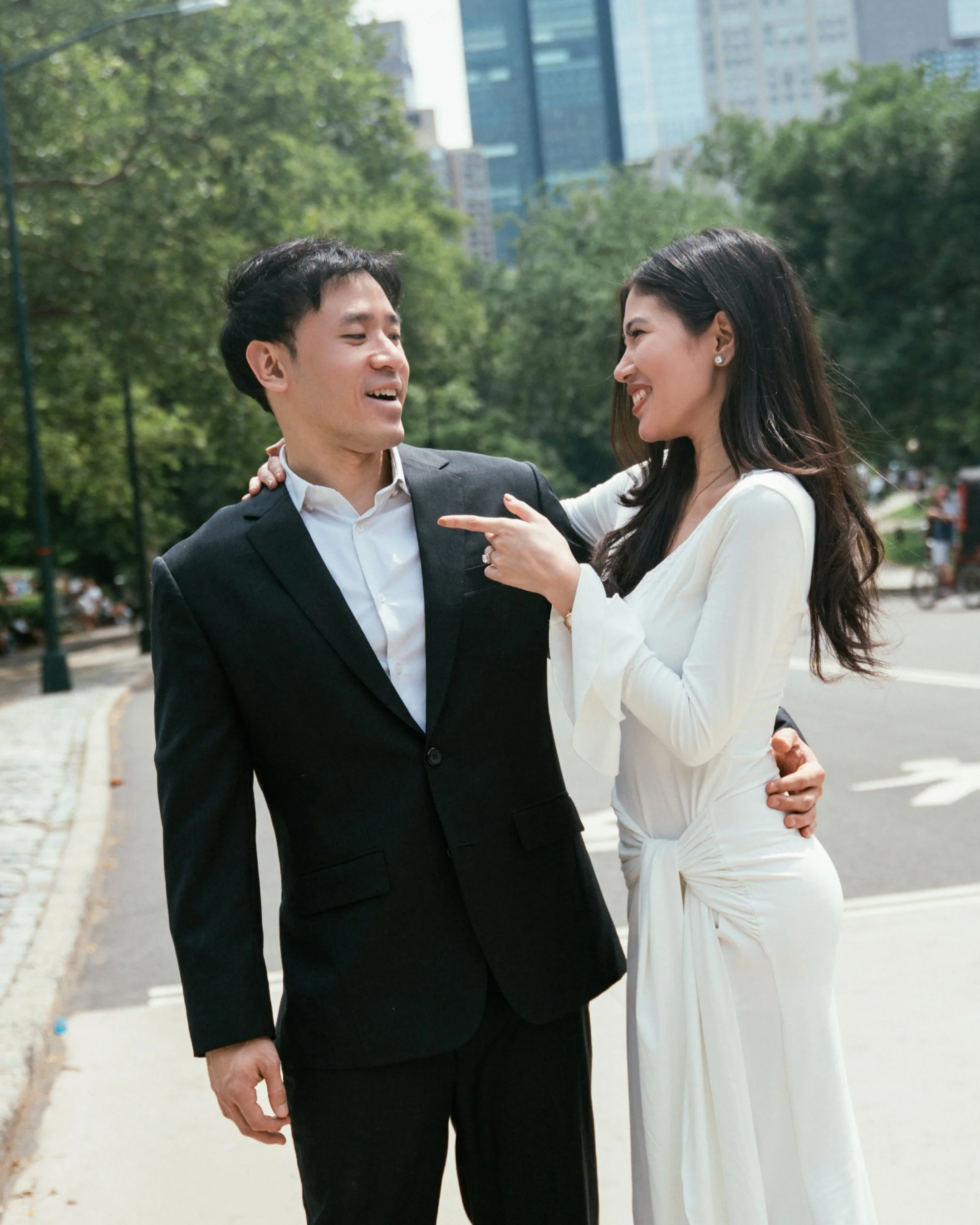 A man and woman are standing outdoors in a city park, smiling and engaging in conversation. The man is wearing a black suit with a white shirt, and the woman is wearing a white dress with long dark hair. The background shows green trees and tall buil