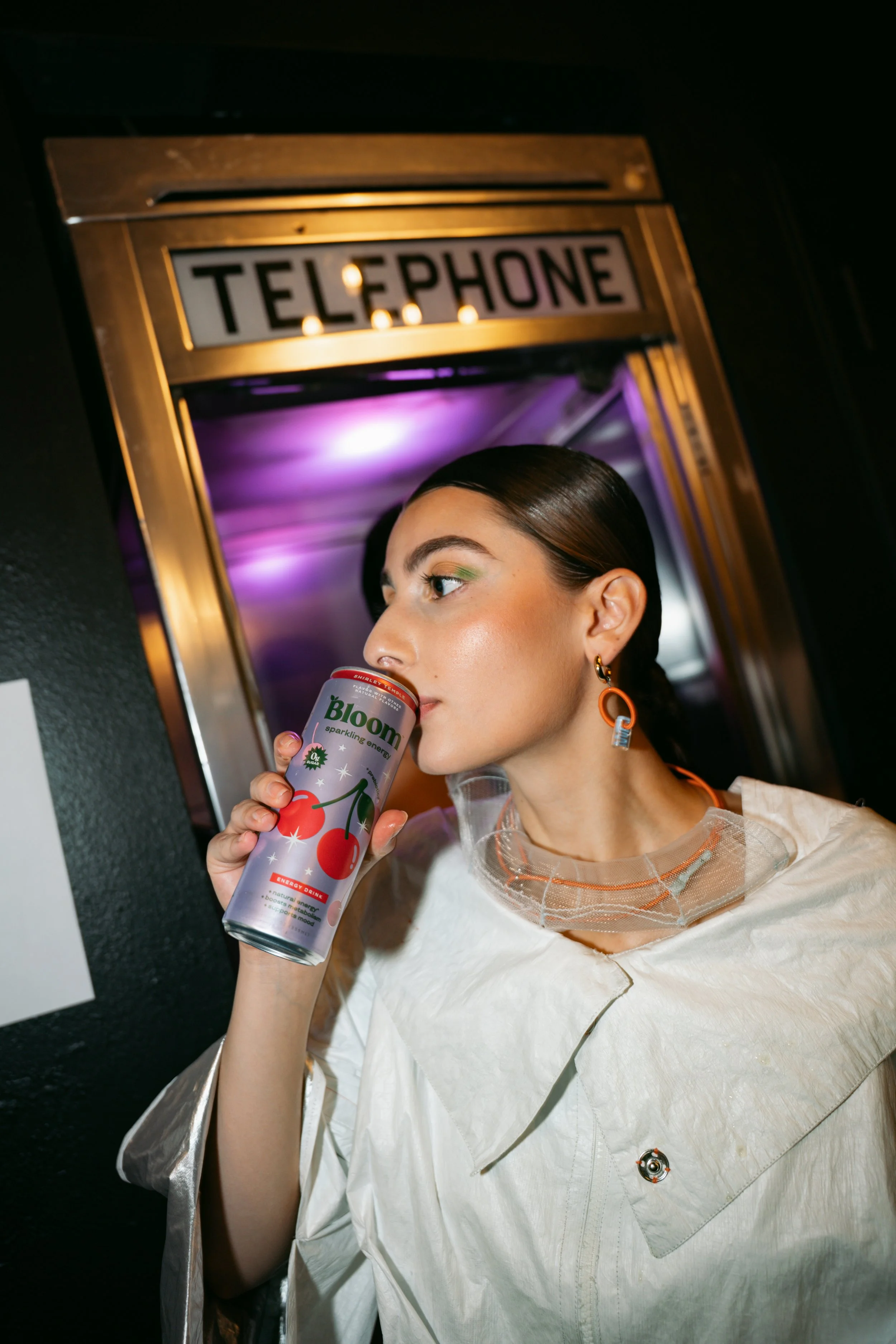 A woman with brown hair and earrings holding a can of Bloom sparkling energy drink. She is standing in front of a mirror with a gold frame labeled 'Telephone,' with purple lighting reflected behind her. She is wearing a white outfit with a transparen