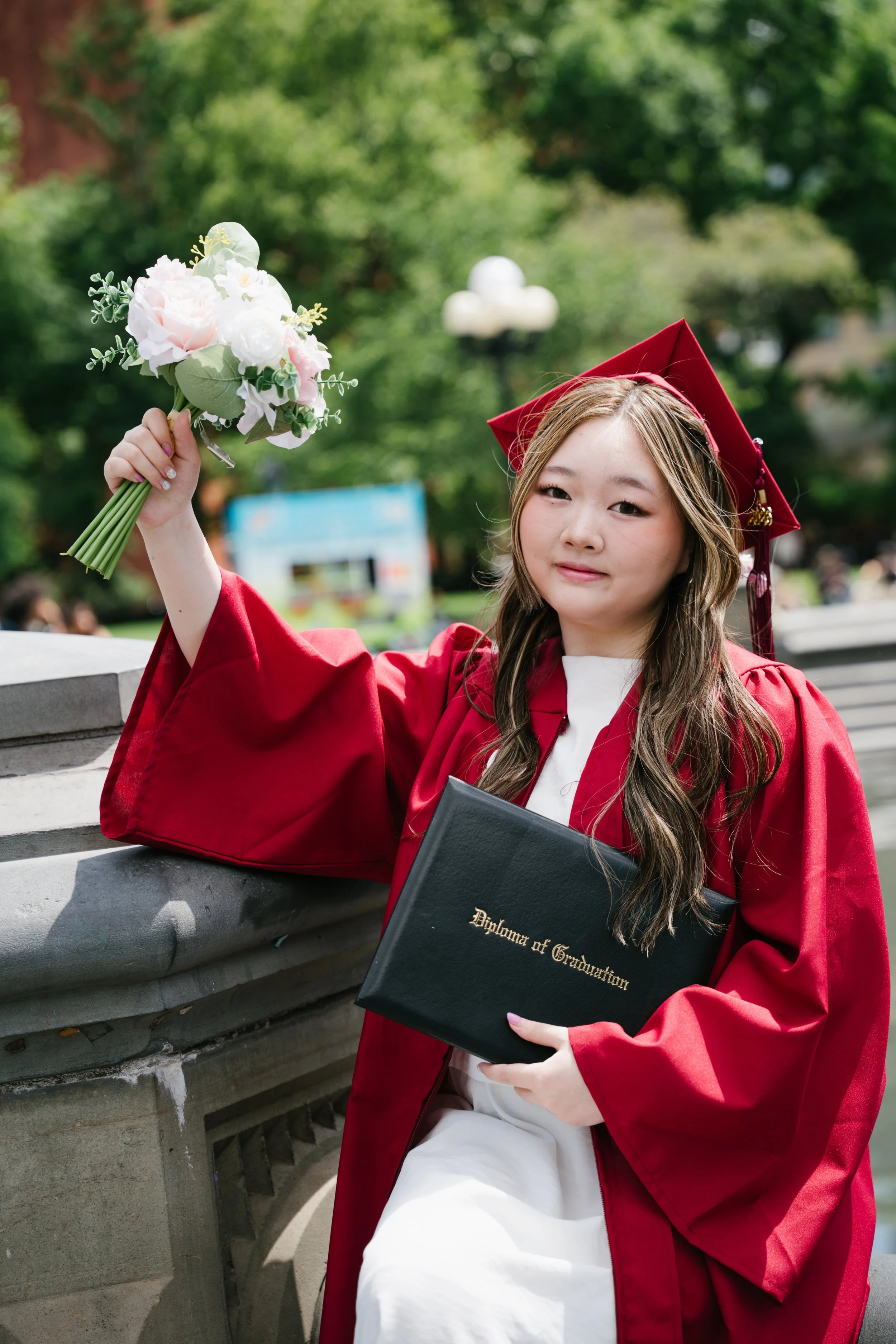 A young woman in a red graduation gown and cap is sitting on a bench outside, holding a bouquet of pink and white flowers in one hand and a black diploma cover with gold lettering in the other, with trees and a lamppost in the background.