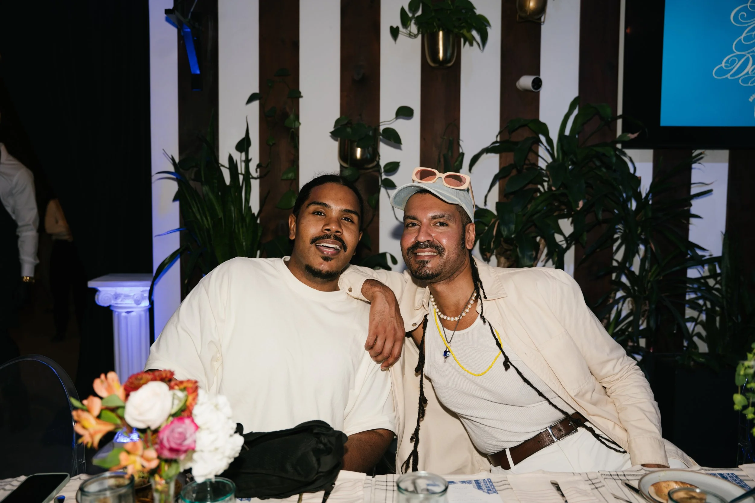 Two men sitting at a table with a floral centerpiece, smiling. They are dressed in light-colored shirts, one wearing a cap with sunglasses on top, the other with braided hair. The background includes green plants and a wooden panel wall, with part of