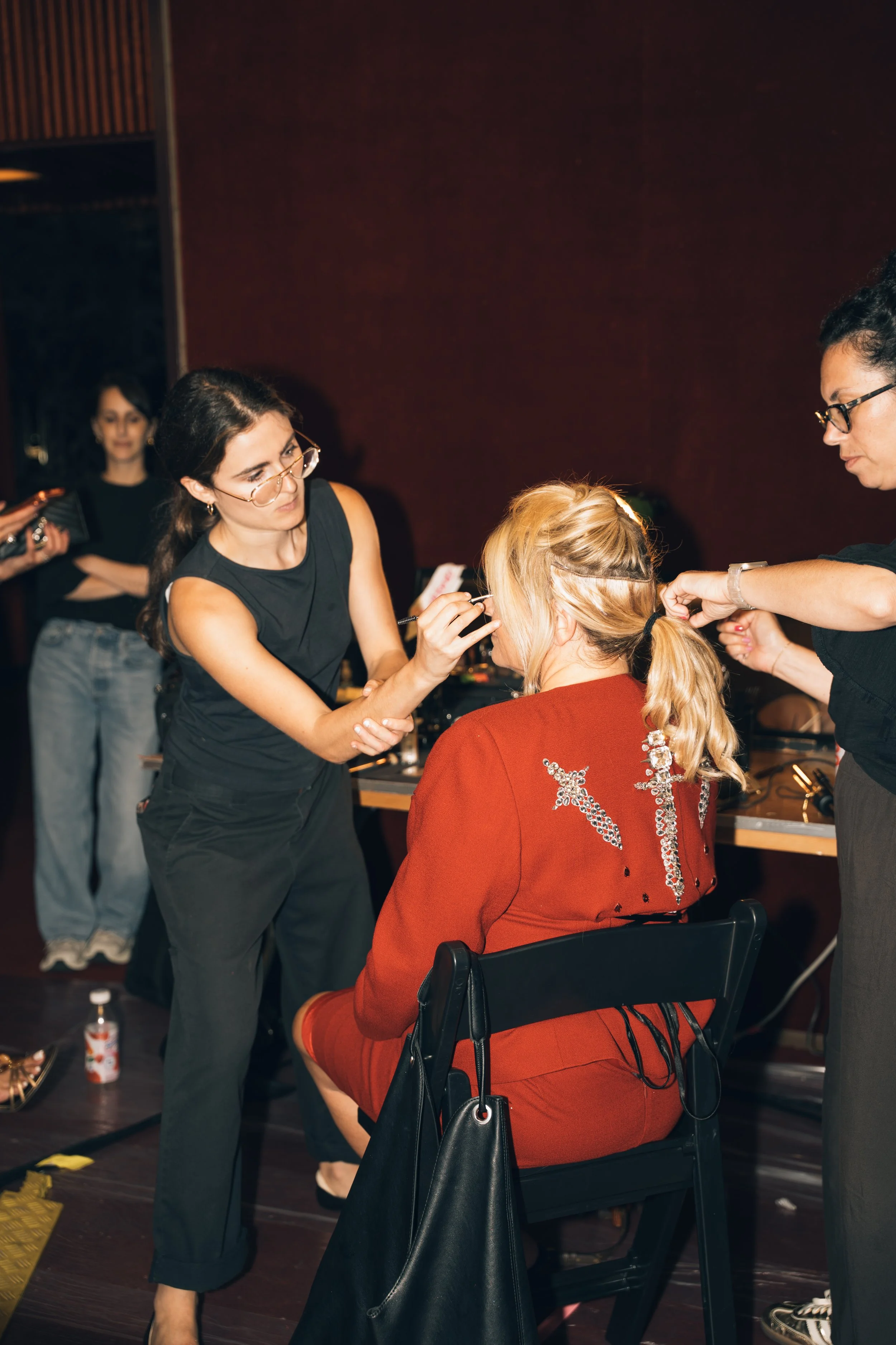 A woman in a red sweater sitting in a chair, getting her makeup done by a makeup artist in a black sleeveless shirt, while another woman in glasses watches. A third person in the background holds a notebook.
