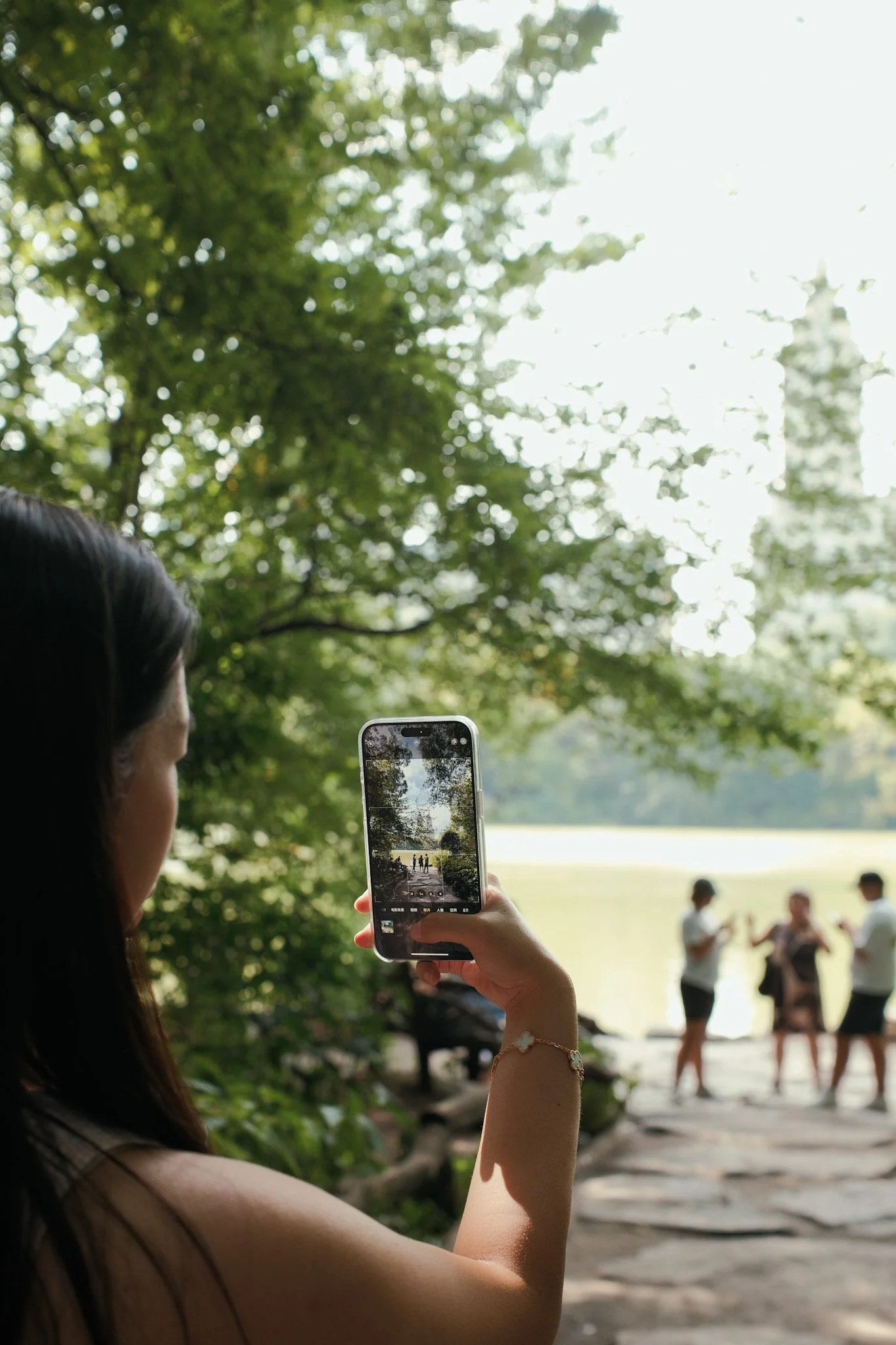 A woman with long dark hair using a smartphone to take a photo in a park with trees and a lake in the background. People are visible in the distance, some taking photos or talking.