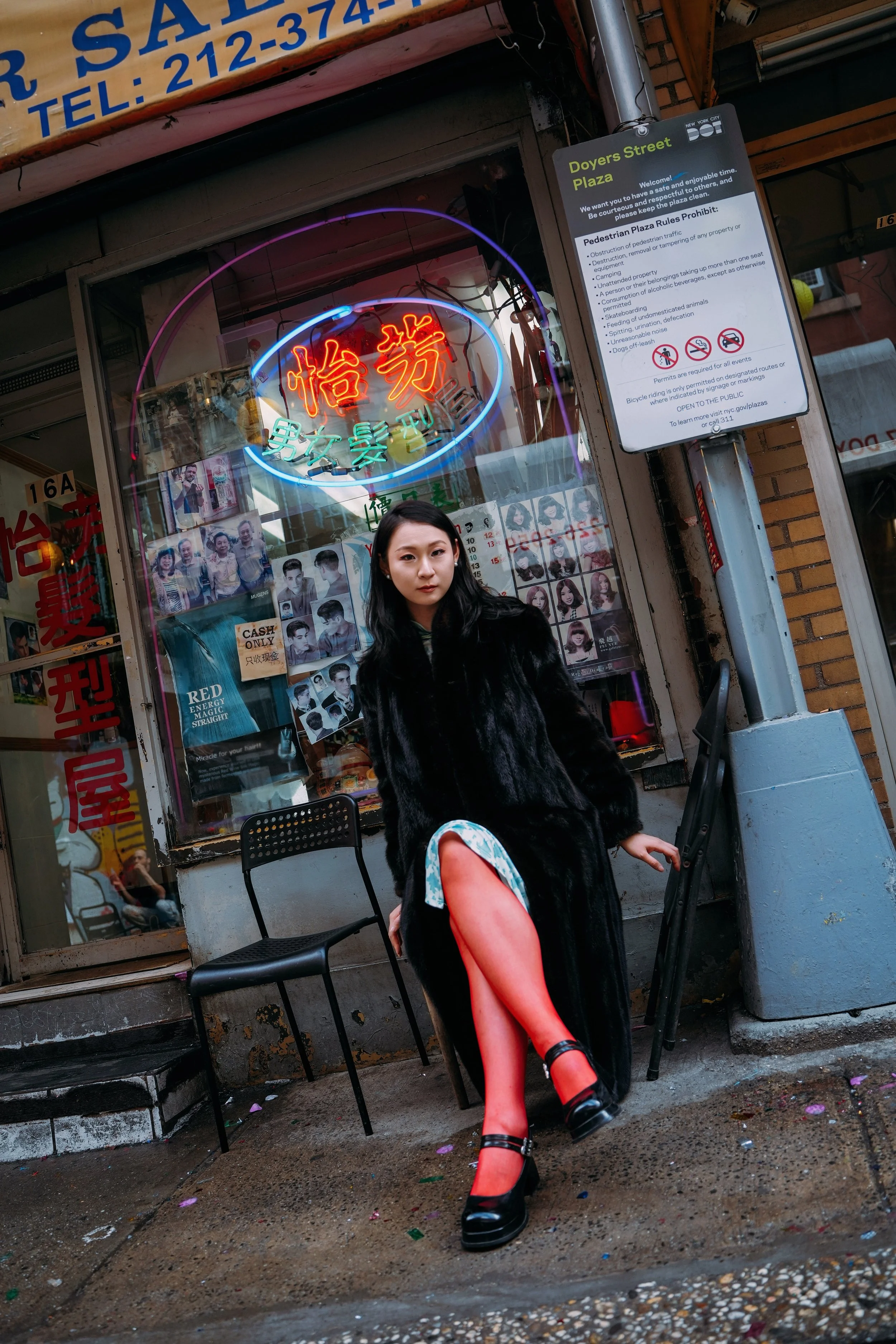 A young woman with black hair, wearing a black fur coat, bright red stockings, and black platform shoes, is sitting outside on a black chair in front of a storefront with neon signs and a sign with rules and regulations. She is looking at the camera with a neutral expression, and her right hand is resting on the chair.