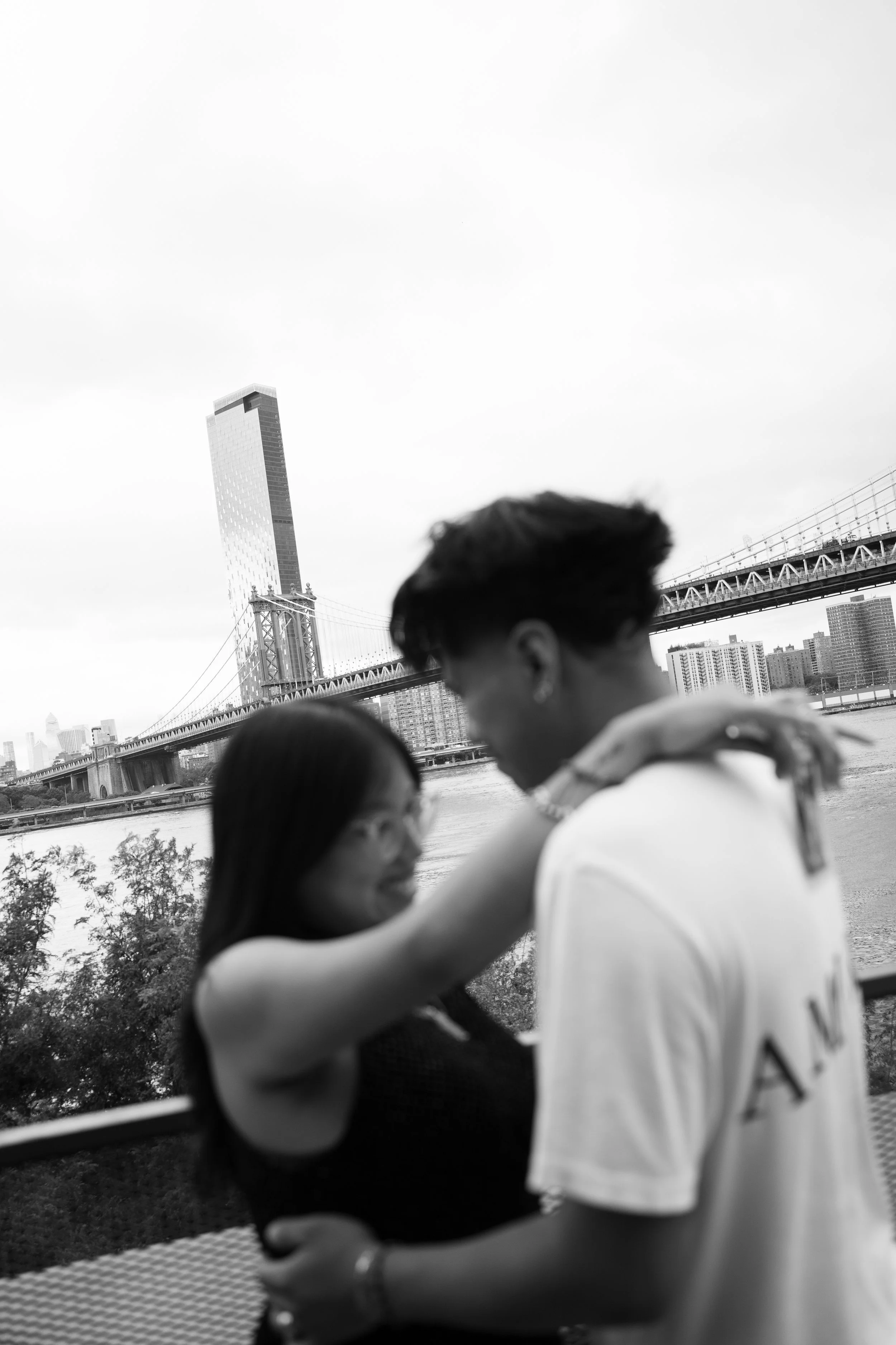 Two people, a man and a woman, holding each other and smiling with the Brooklyn Bridge and city skyline in the background.