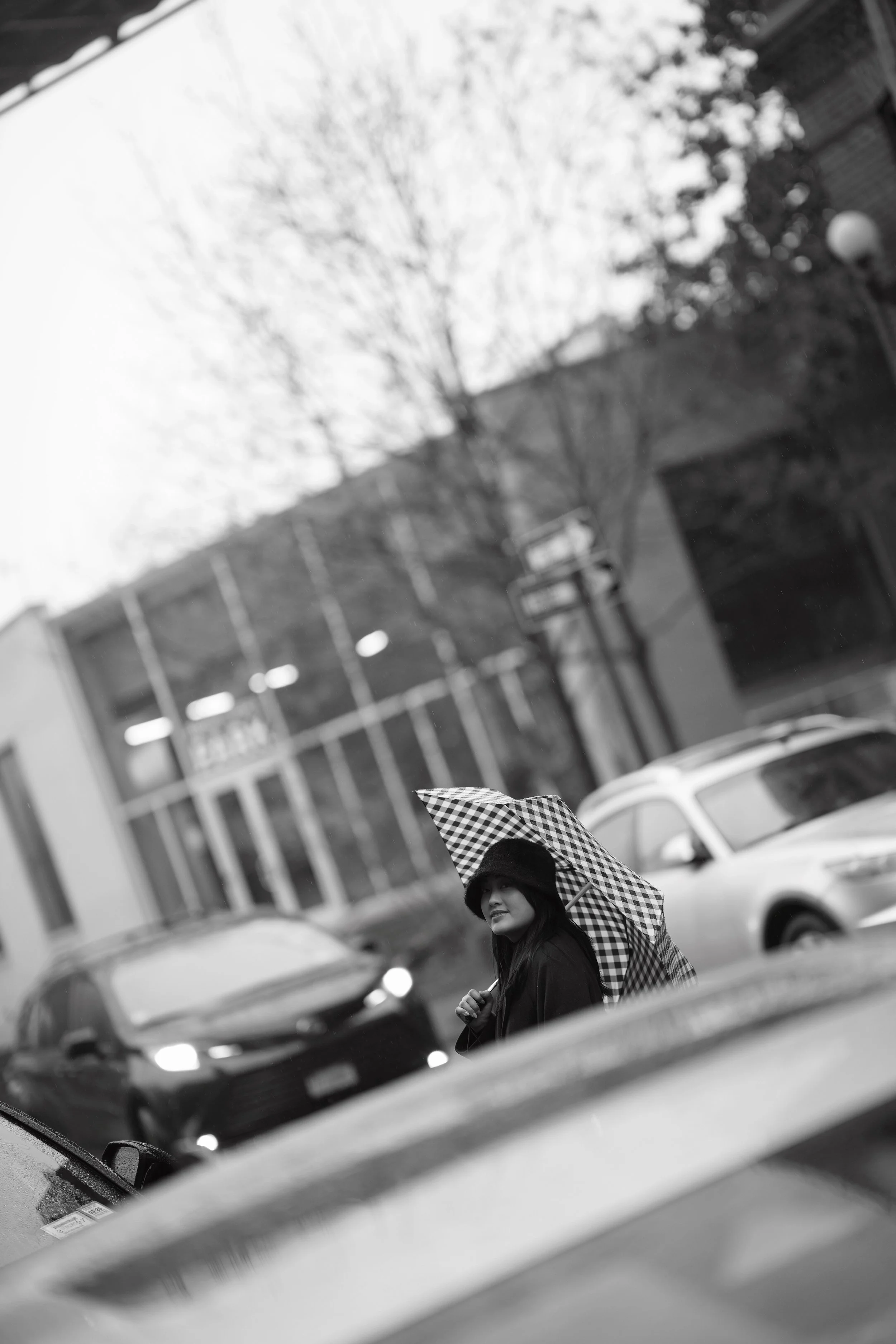 A woman with long hair and a hat holding a checkered umbrella walking outdoors on a rainy day.
